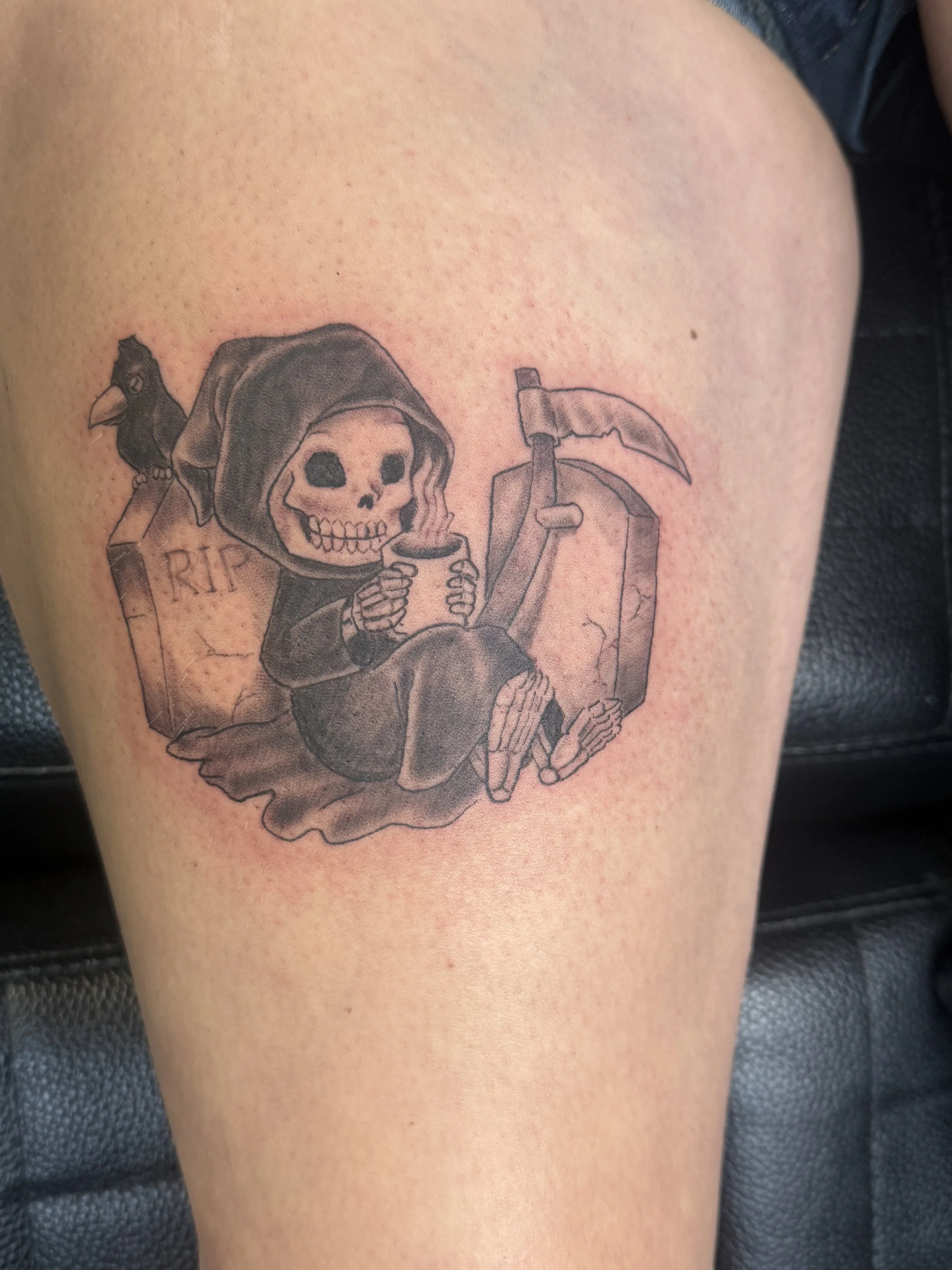 Tattoo of a skeleton with a hoodie, sitting and holding a mug, next to a tombstone with 'RIP' written on it, and holding a scythe, with a black bird perched on the tombstone.