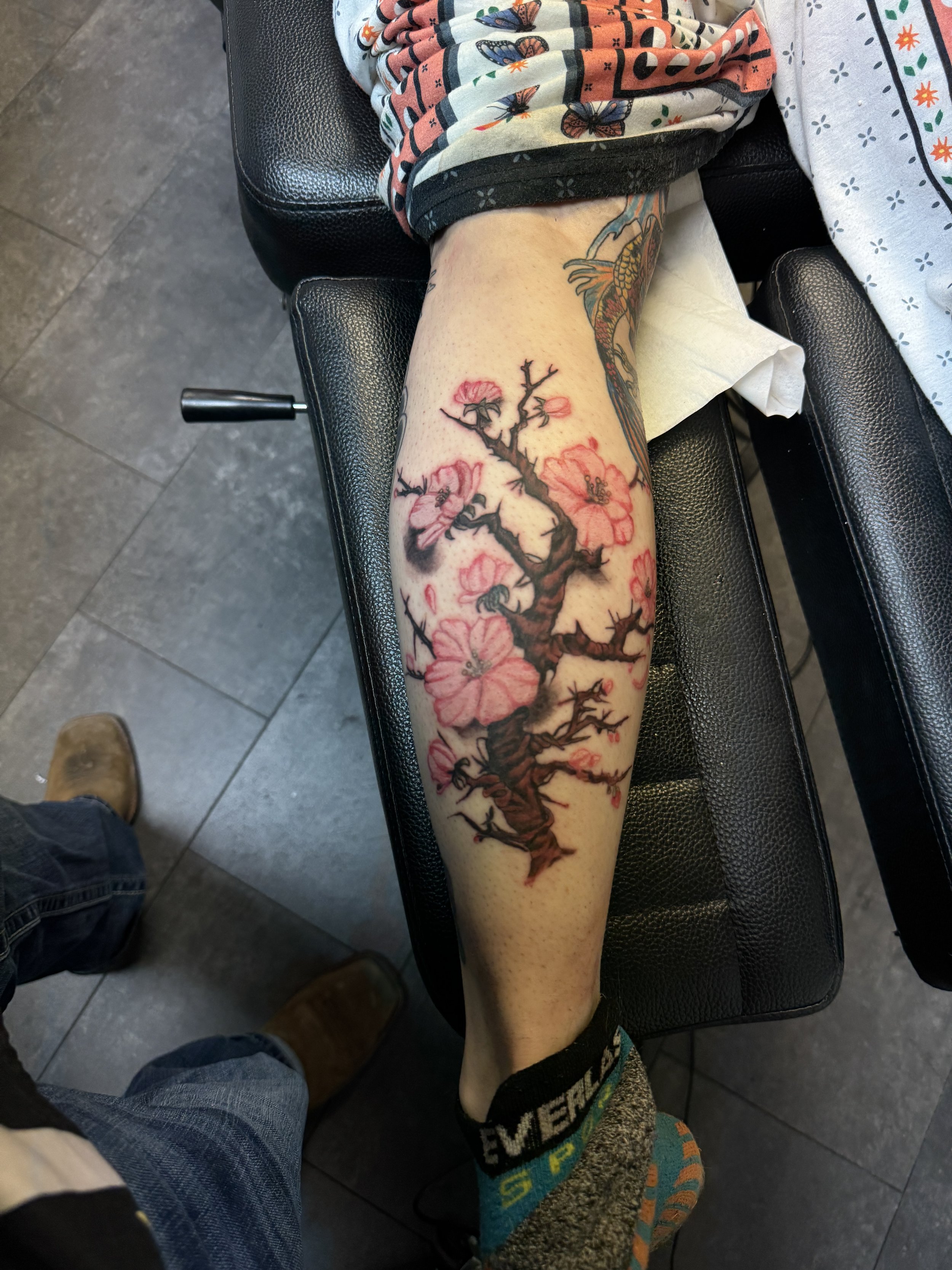 A person's leg with a colorful tattoo of a cherry blossom tree with pink flowers and dark brown branches on their calf.