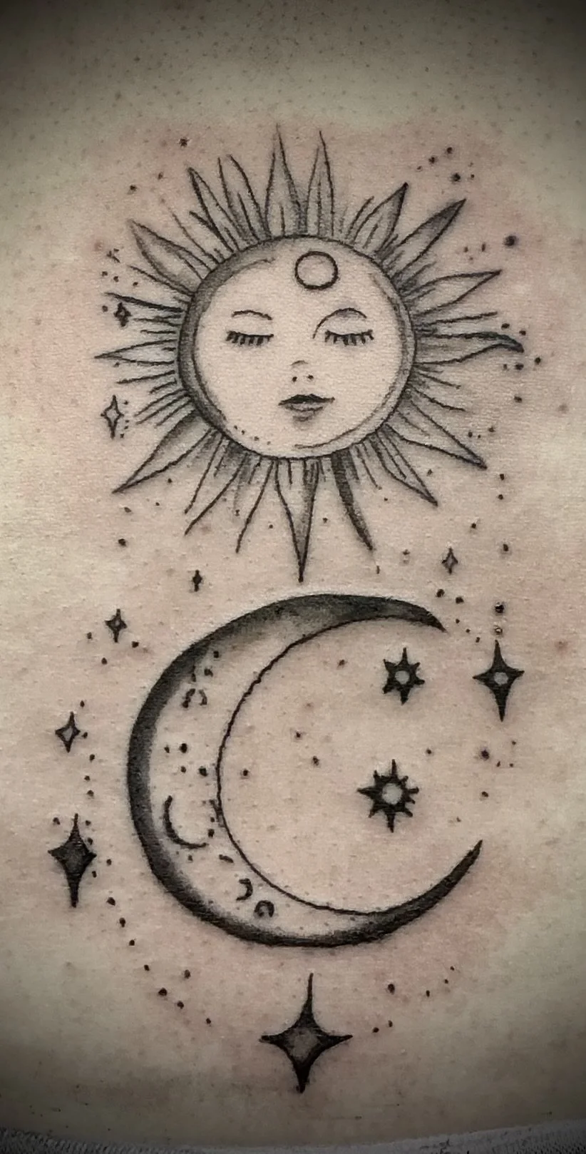 Tattoo of a moon with a face, surrounded by stars, and a sun with a face at the top. The moon has closed eyes, and the sun has a calm expression.