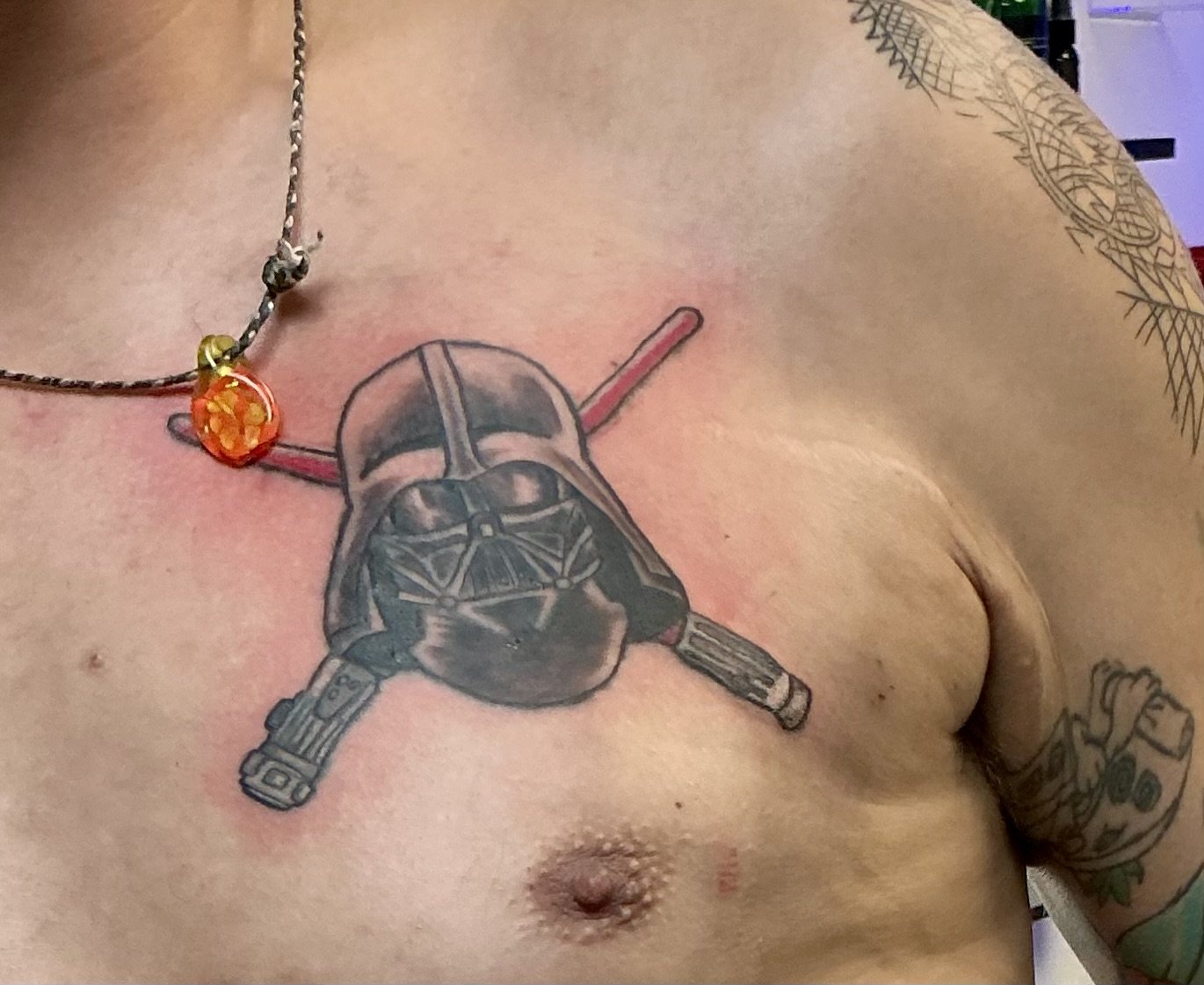 Tattoo of a Darth Vader helmet with crossed lightsabers on the chest of a person with tattoos on the arm and necklaces around the neck.