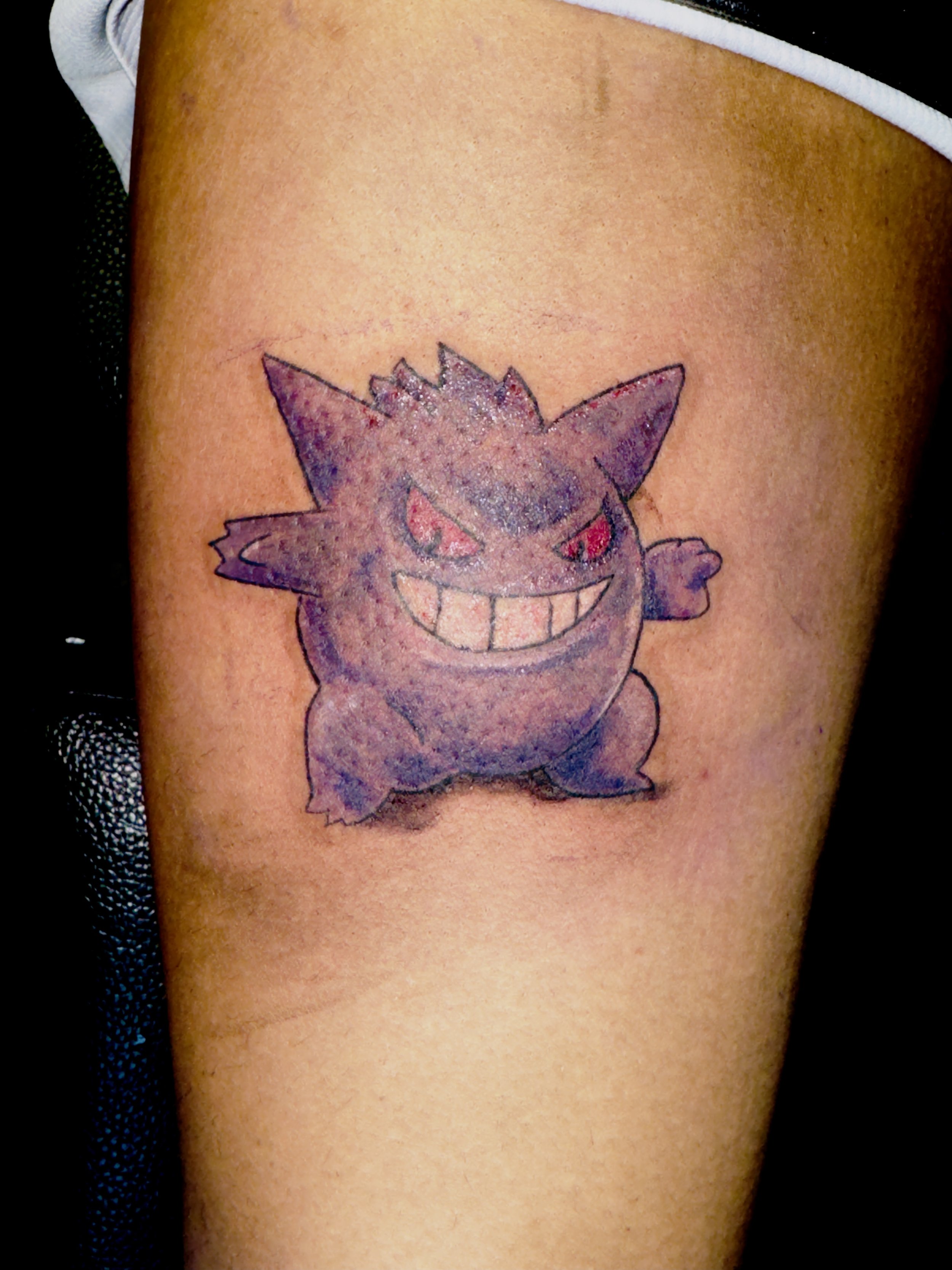 Tattoo of Gengar from Pokémon on person's upper arm, smiling with red eyes and sharp teeth.