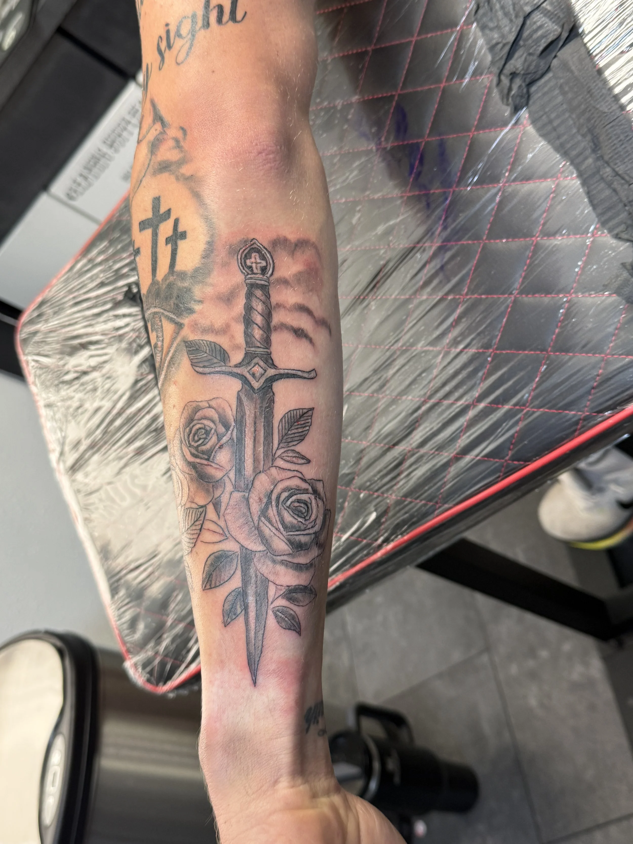 Tattoo of a sword with roses and leaves on a person's forearm.