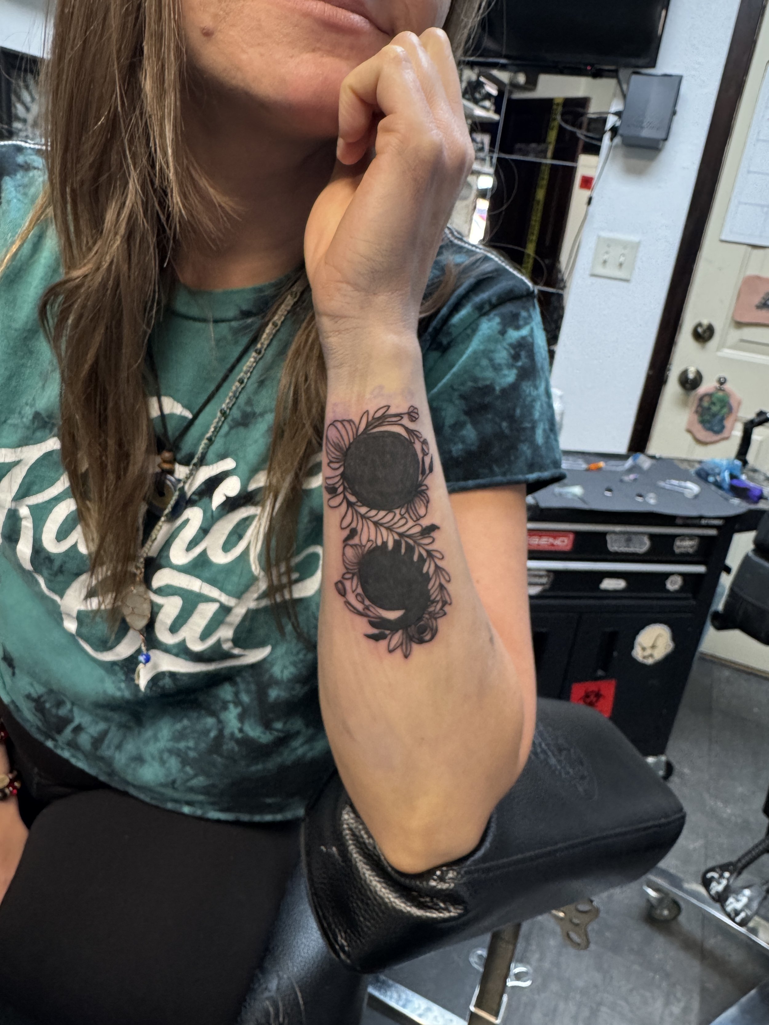 A woman with a tattoo of two circular shapes surrounded by floral and leaf designs on her forearm, seated in a tattoo studio.