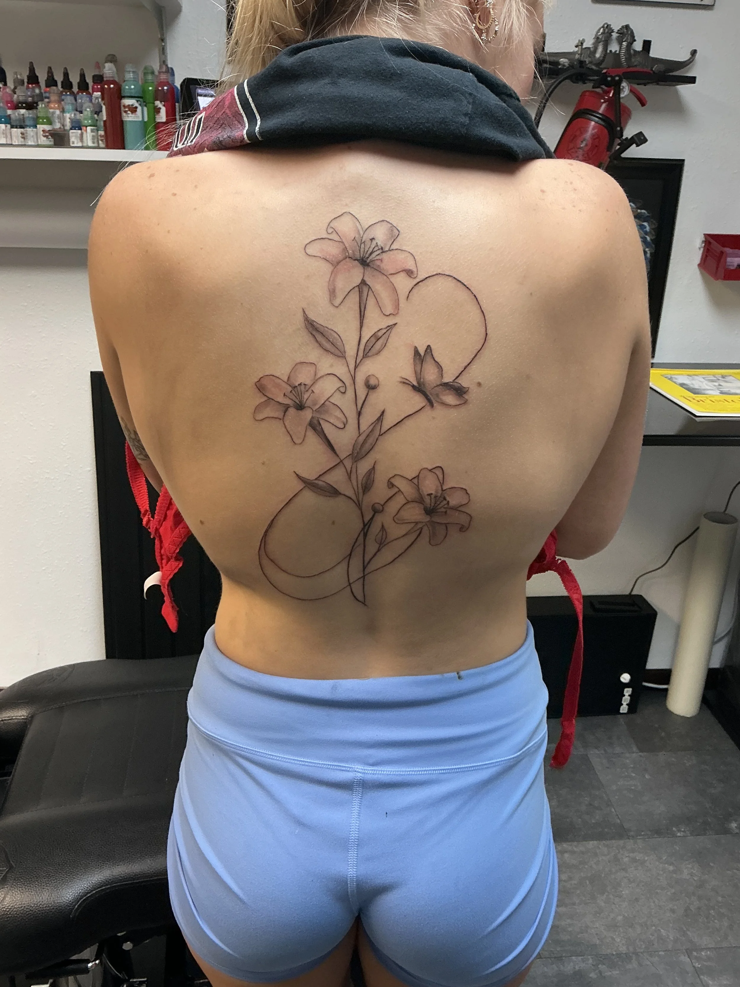 Back of a person with a floral tattoo and a butterfly on the upper back. The person is in a tattoo studio, wearing light blue shorts and a dark top draped around the neck.