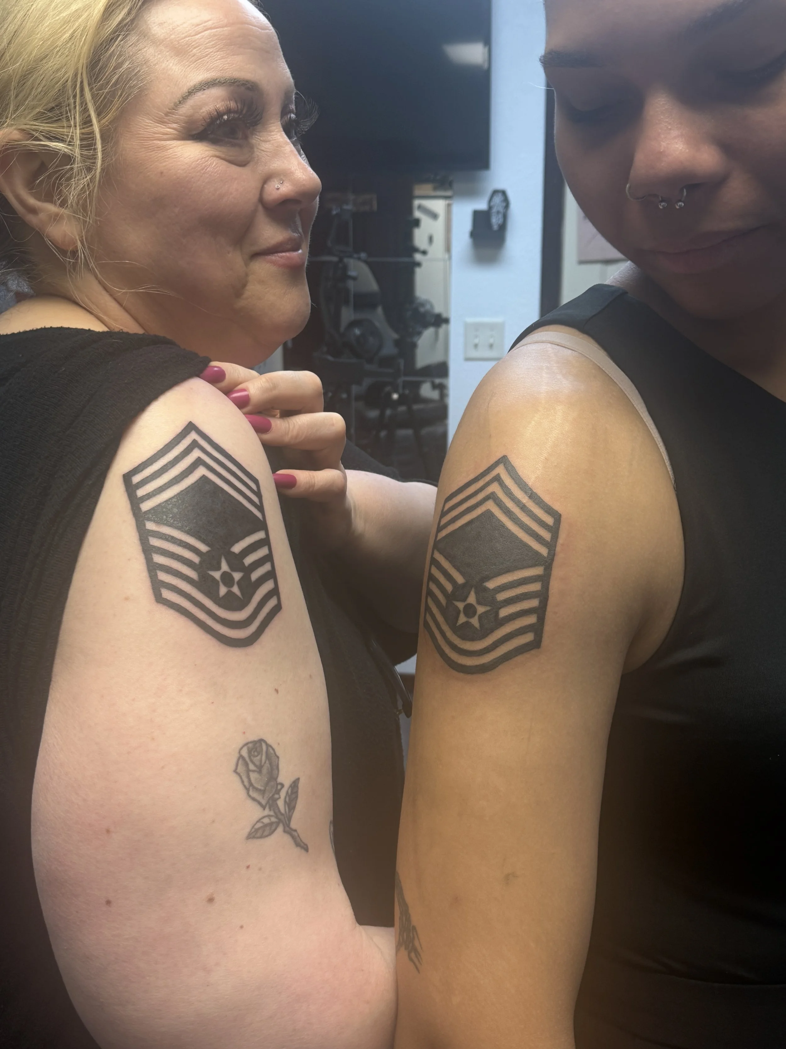 Two women smiling and showing shoulder tattoos with military rank insignia and a rose tattoo on the arm, indoors.