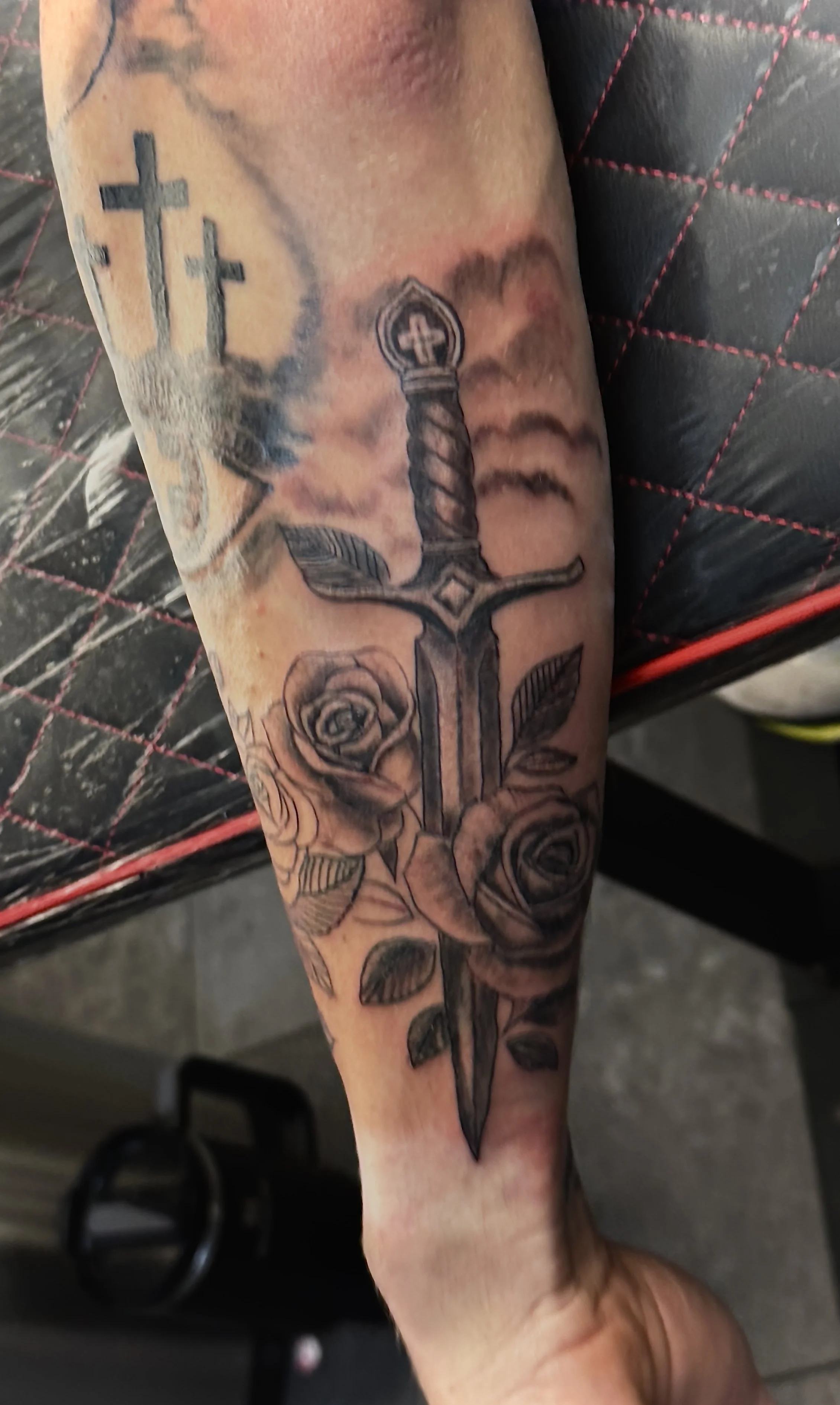 Tattoo of a sword with roses on the forearm, with three crosses in the background.