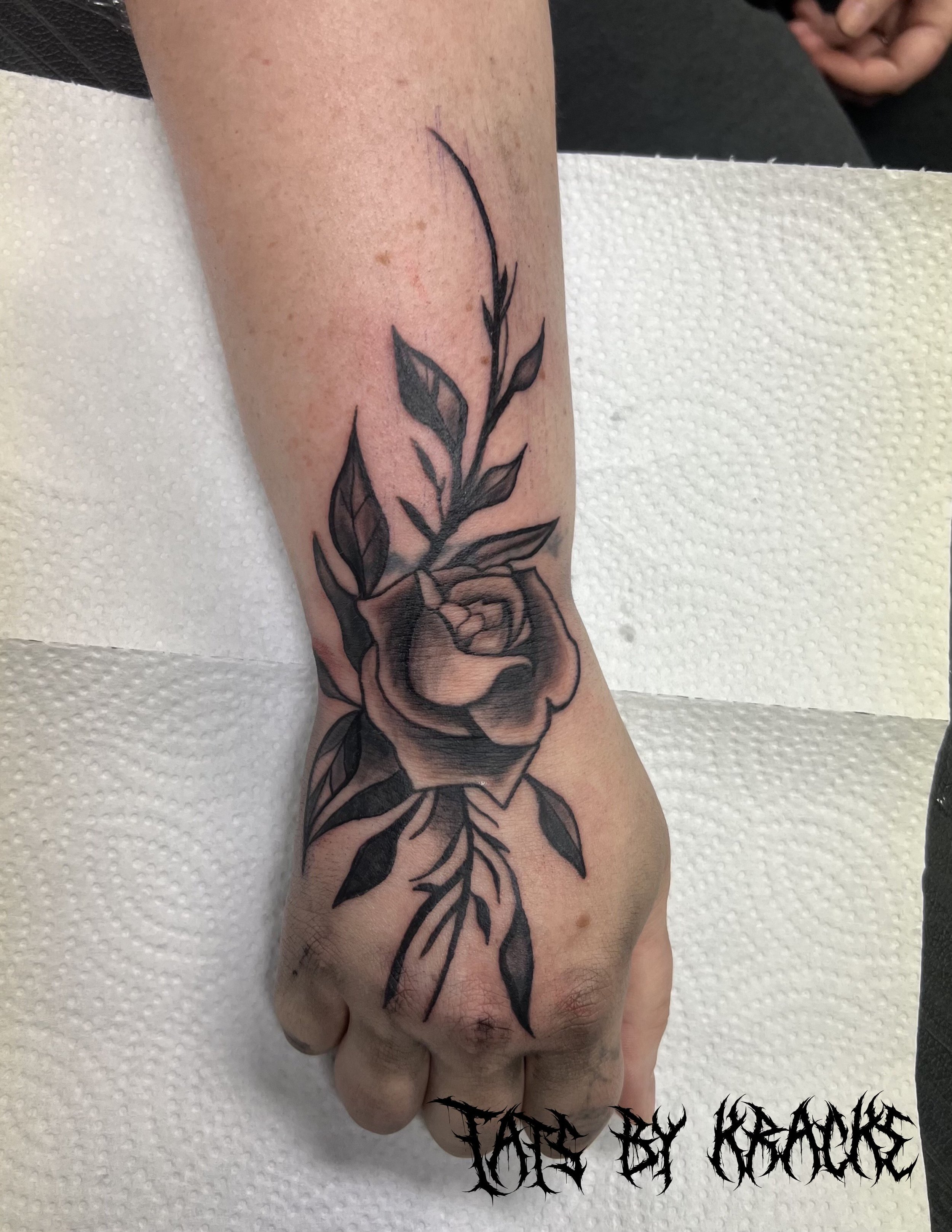 Black and gray tattoo of a rose with leaves on a person's hand and wrist.