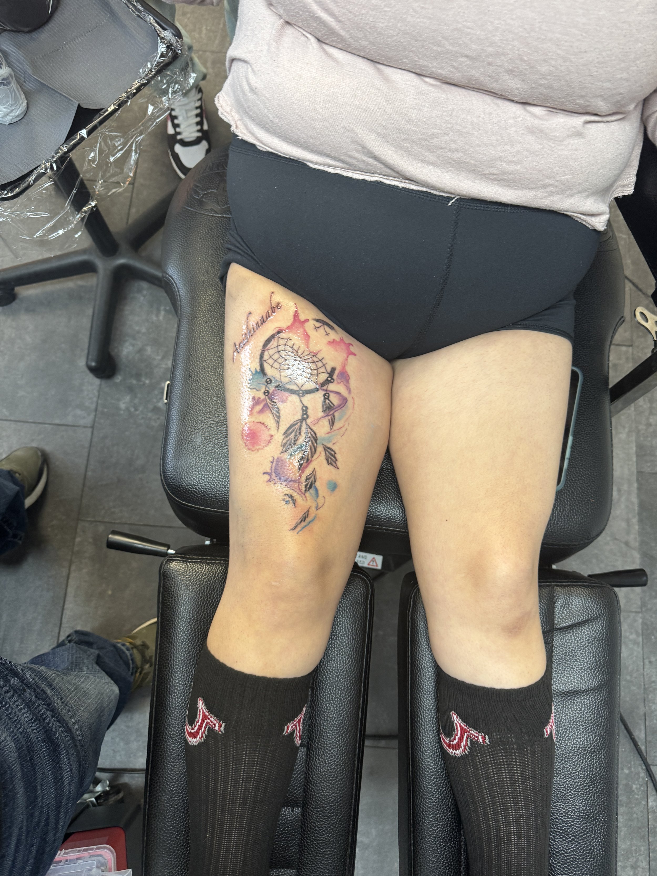 A person sitting on a black tattoo chair with their right thigh visible, showing a colorful tattoo of a dreamcatcher with feathers and stars, and the word 'Animalite' written in cursive. The person is wearing black shorts, a beige T-shirt, black sock