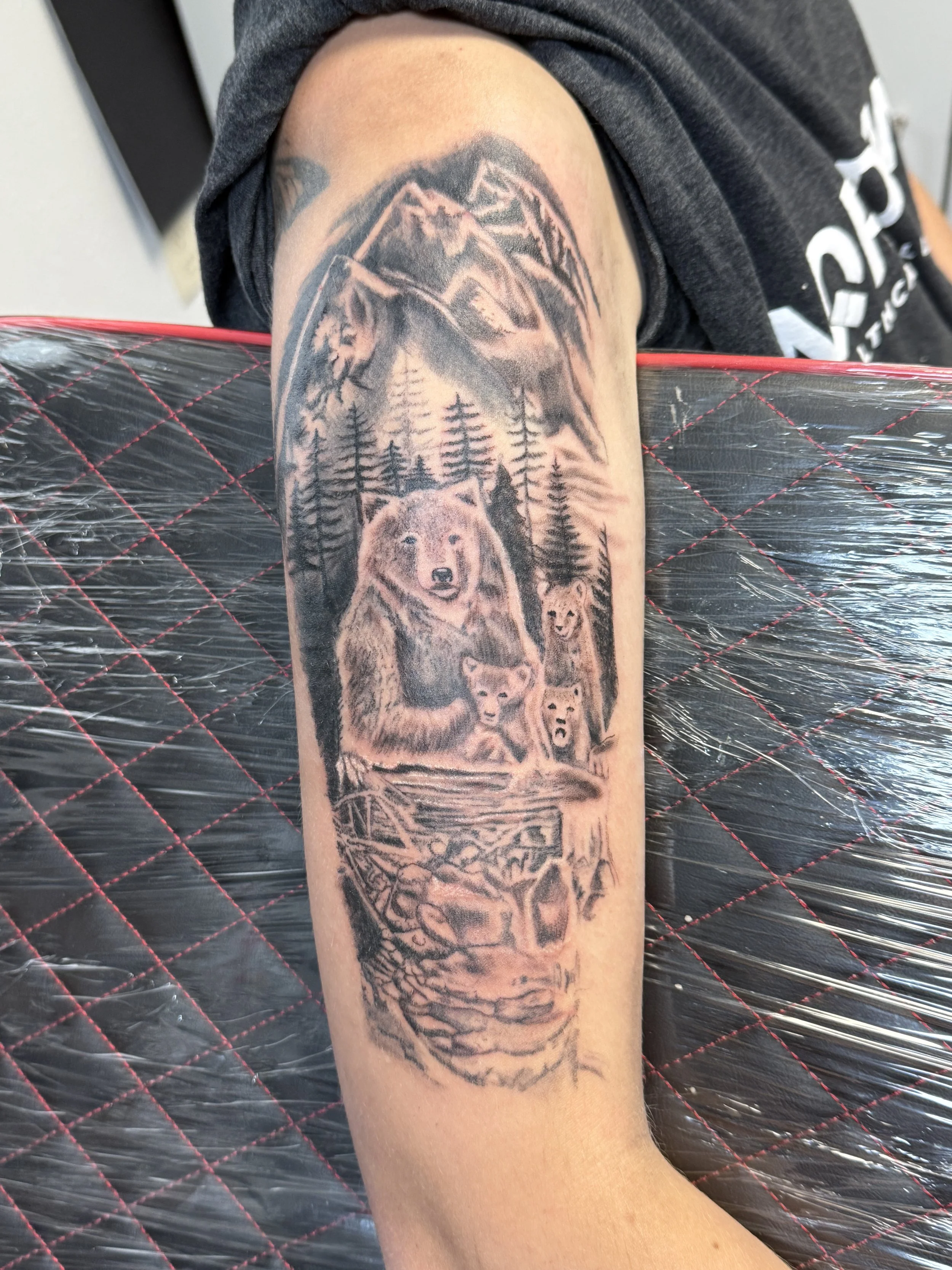 Tattoo of a mountain landscape with a bear, a cub, and a wolf pack, surrounded by pine trees.