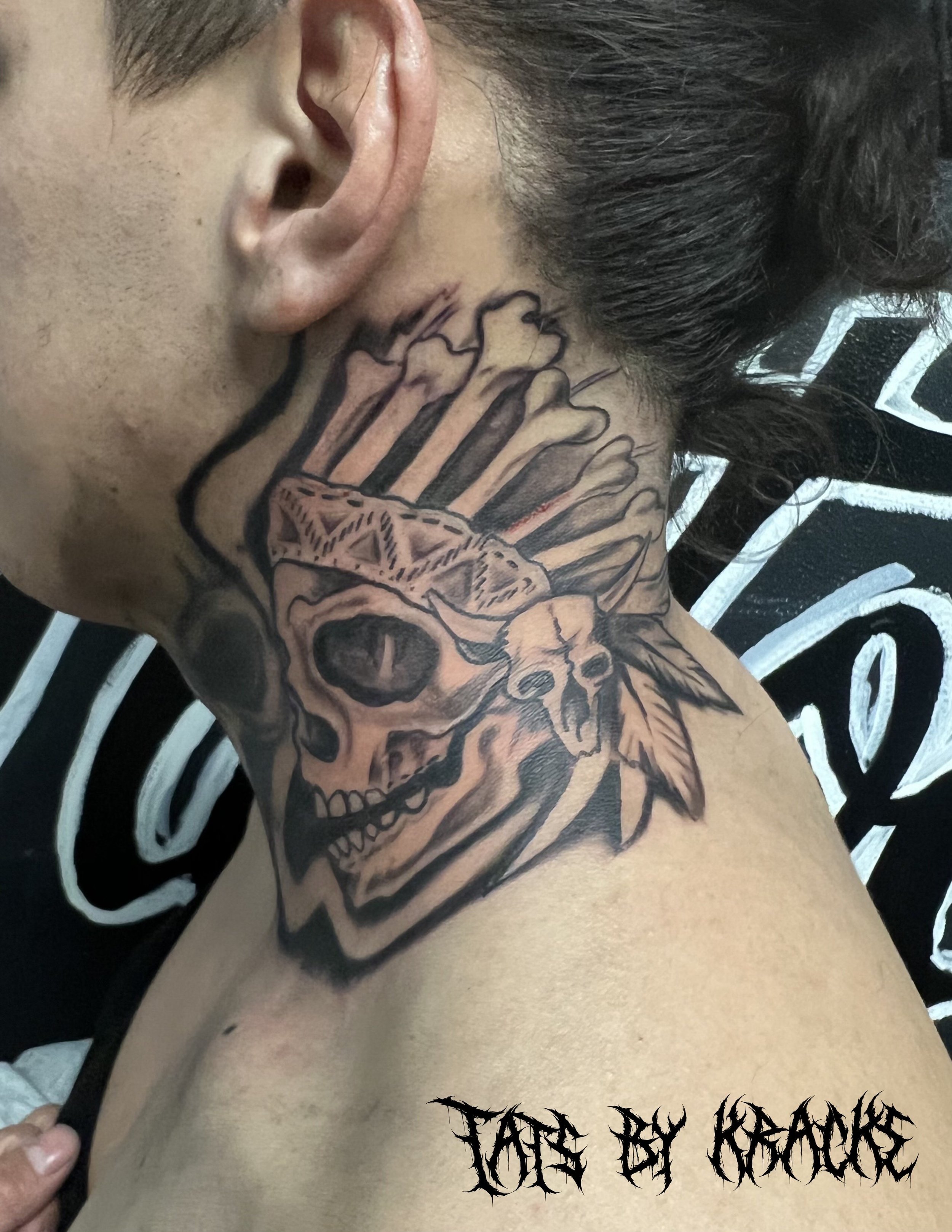 Tattoo on person's neck featuring a skull with feathers and a tribal design with the words 'Tate by KreAKe' in stylized font at the bottom.