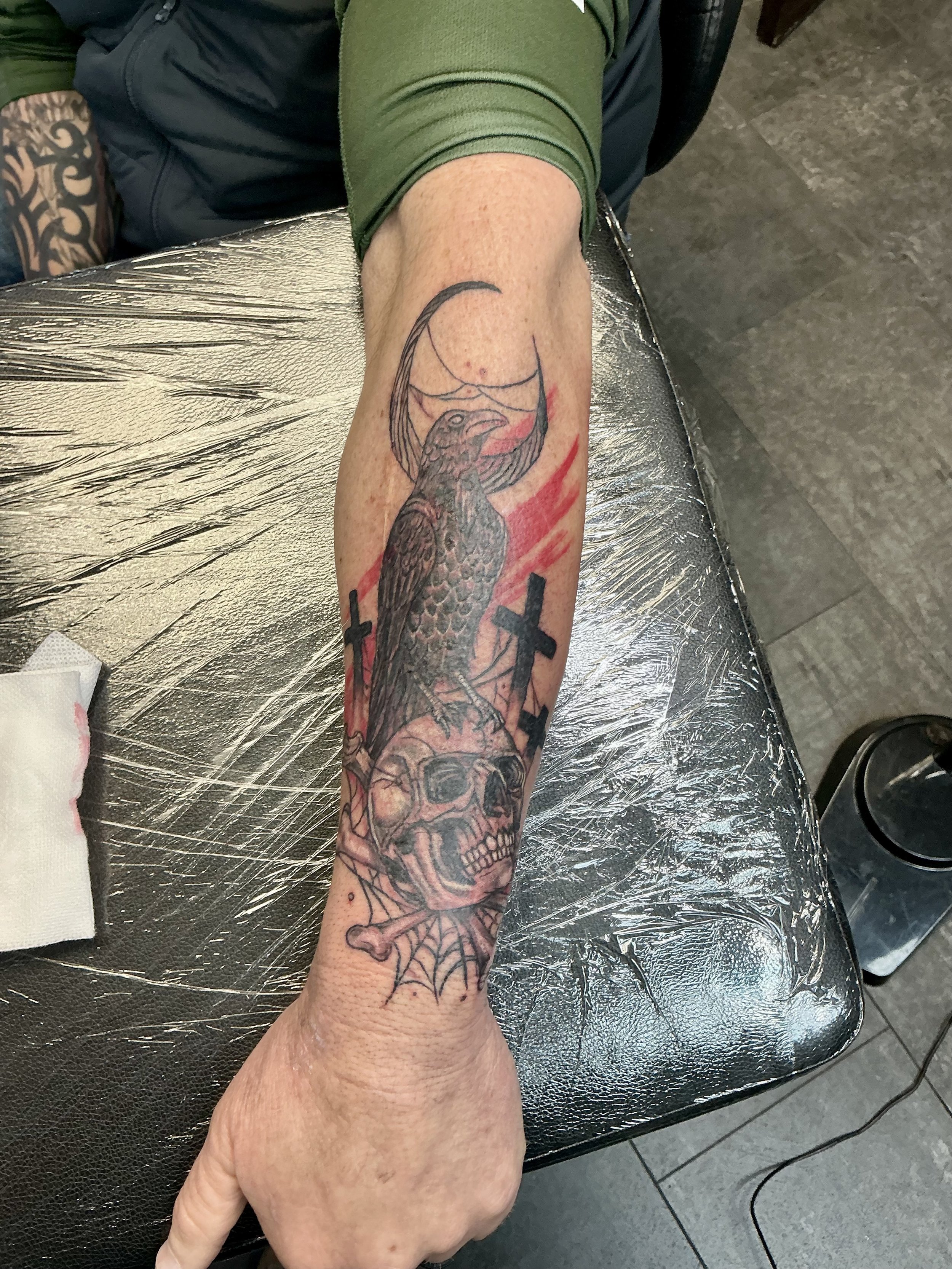 Tattoo of a crow with a crescent moon, two crosses, a human skull with spider web details, and red streaks on a person's forearm.