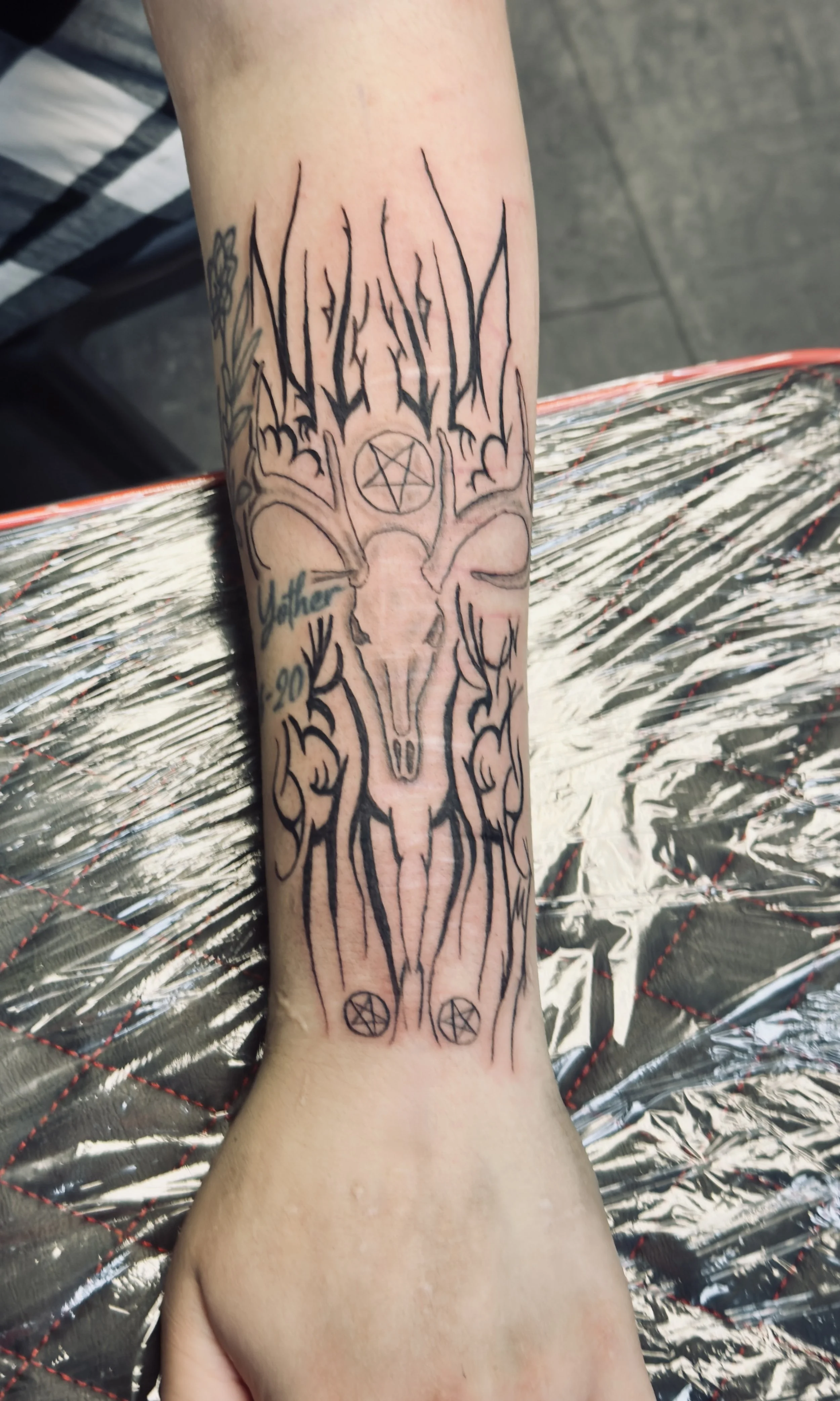 A tattoo on a person's forearm featuring a deer skull with antlers, a pentagram, and the word 'Mother 90' in blue ink.