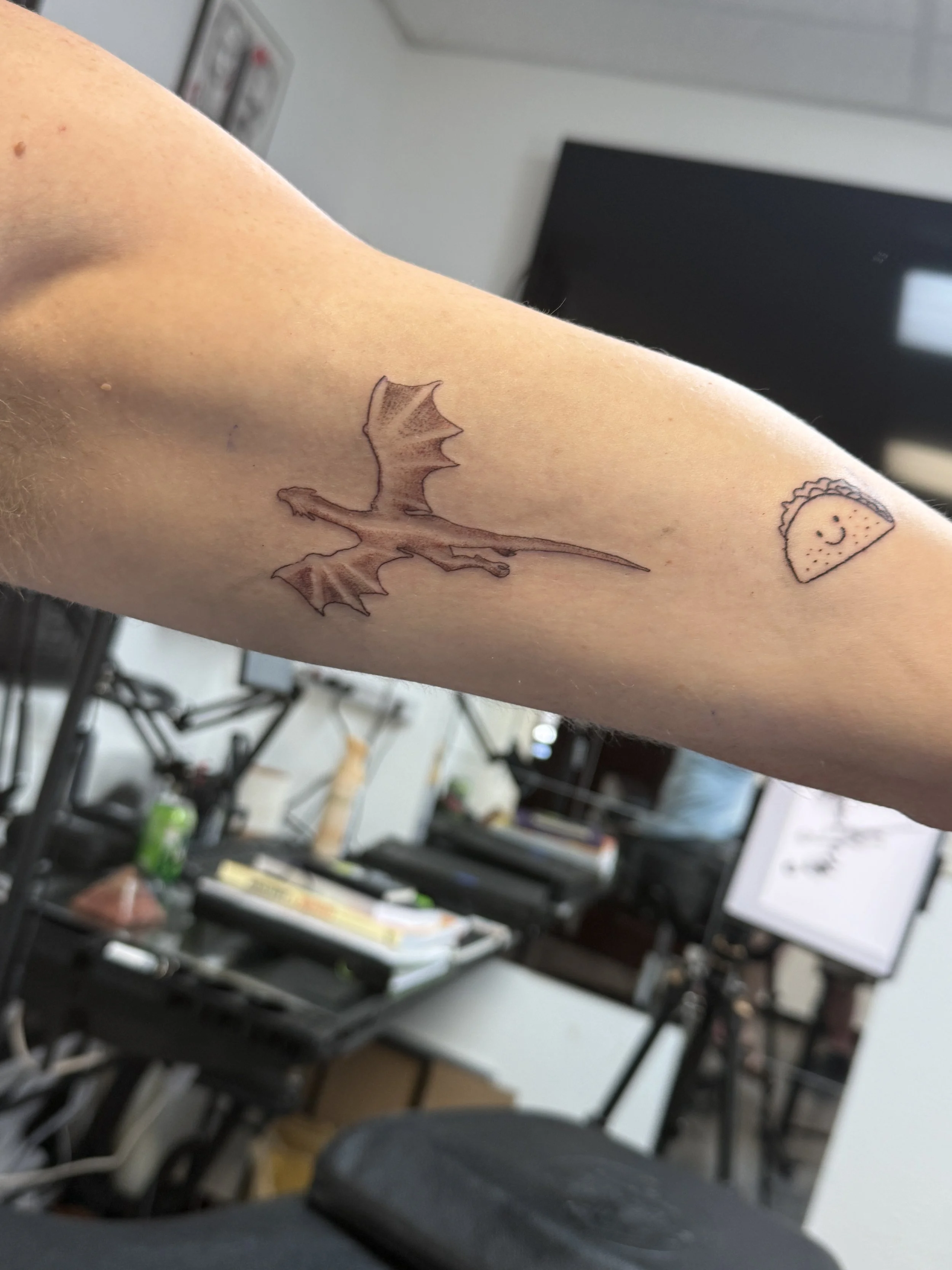 Tattoo of a pterodactyl flying on a person's forearm, next to a small taco with a smiling face.