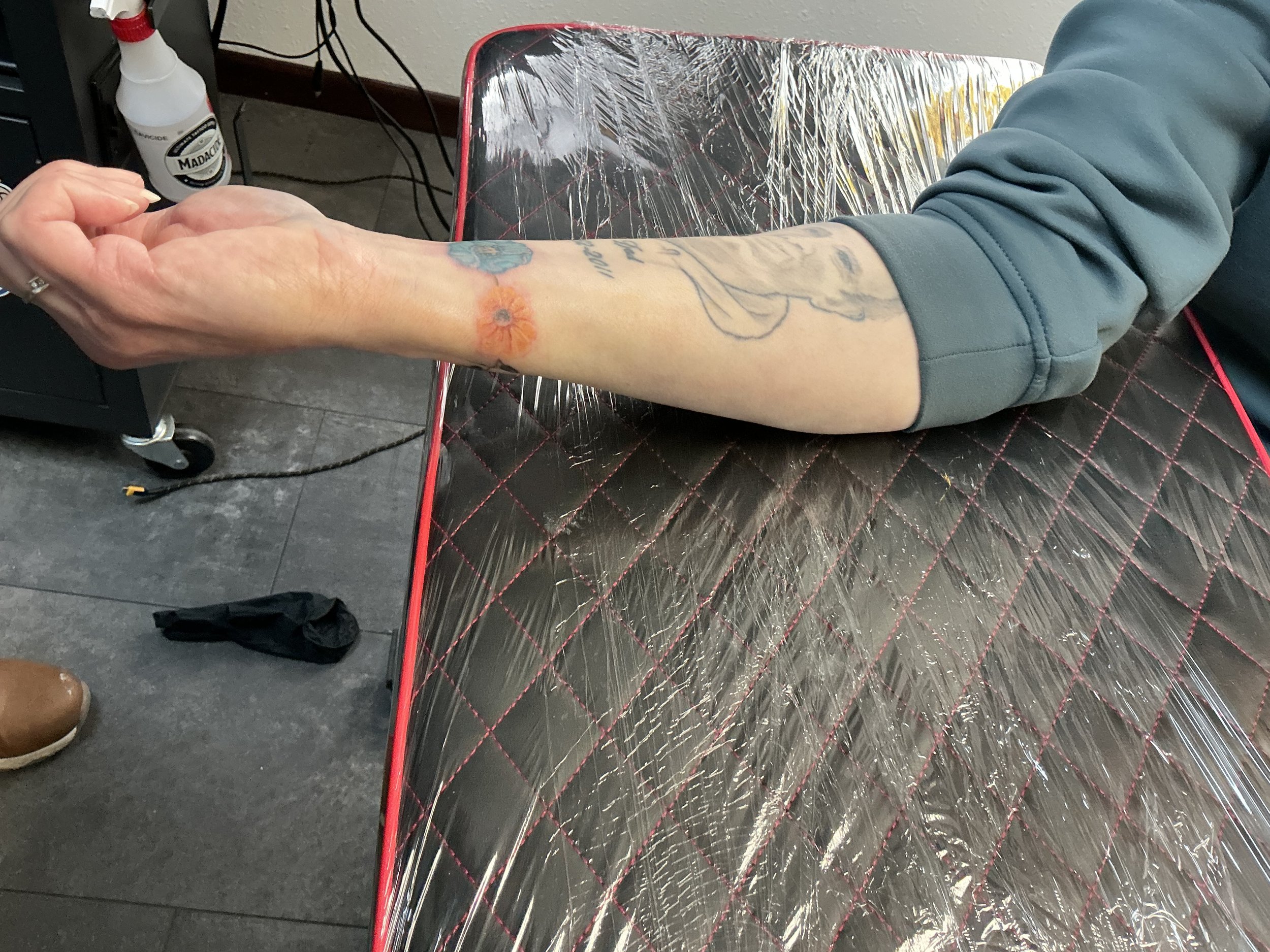 A person's arm resting on a tattoo table with a black and red covering, showing tattoos including a colorful flower and an outline of a face.