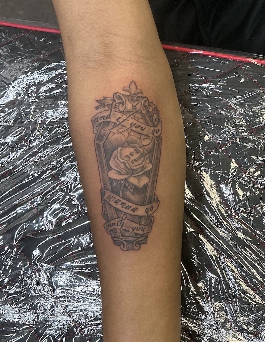 Tattoo of a coffin with a rose inside, surrounded by a decorative frame, with a ribbon banner reading 'Loading with you'.