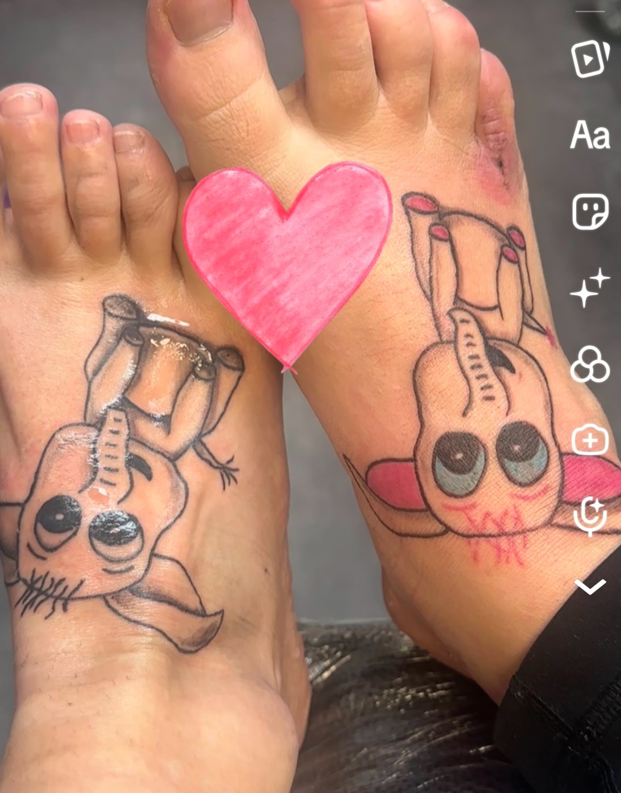 Two hands with matching tattoos of cartoon-like animals, upside down, with a pink heart overlaying the center.