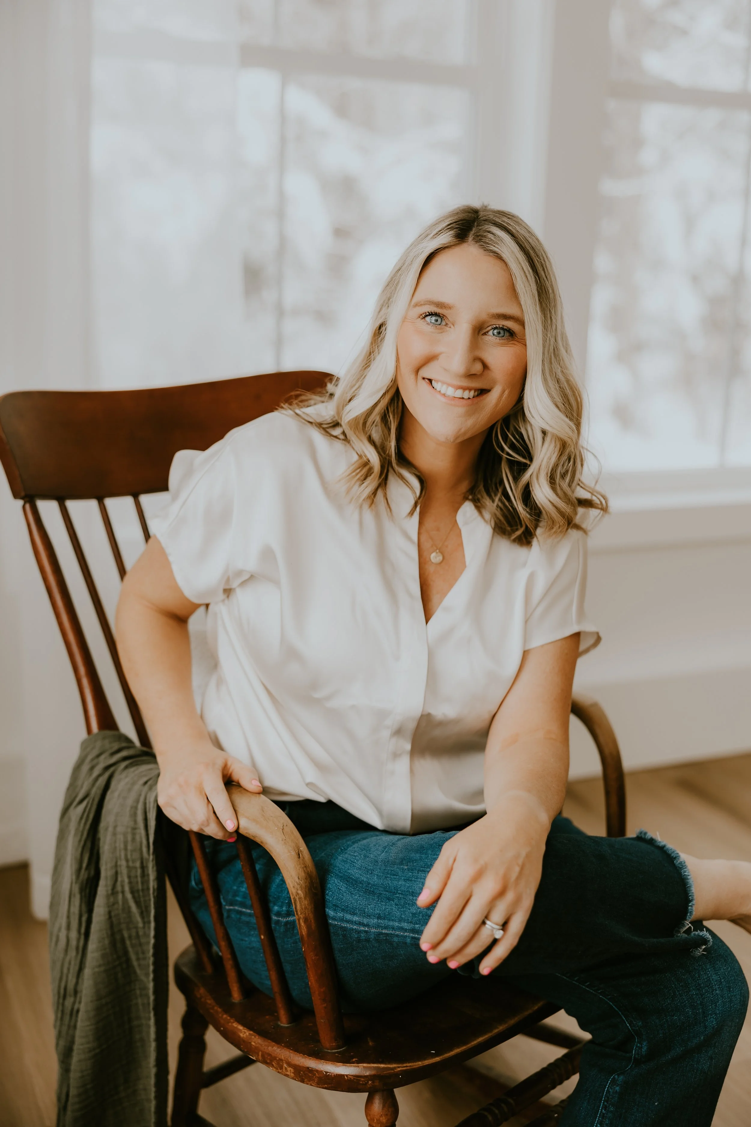 A woman with blonde hair and blue eyes sitting on a wooden chair, smiling at the camera, wearing a white shirt and jeans, in a room with large windows and natural light.