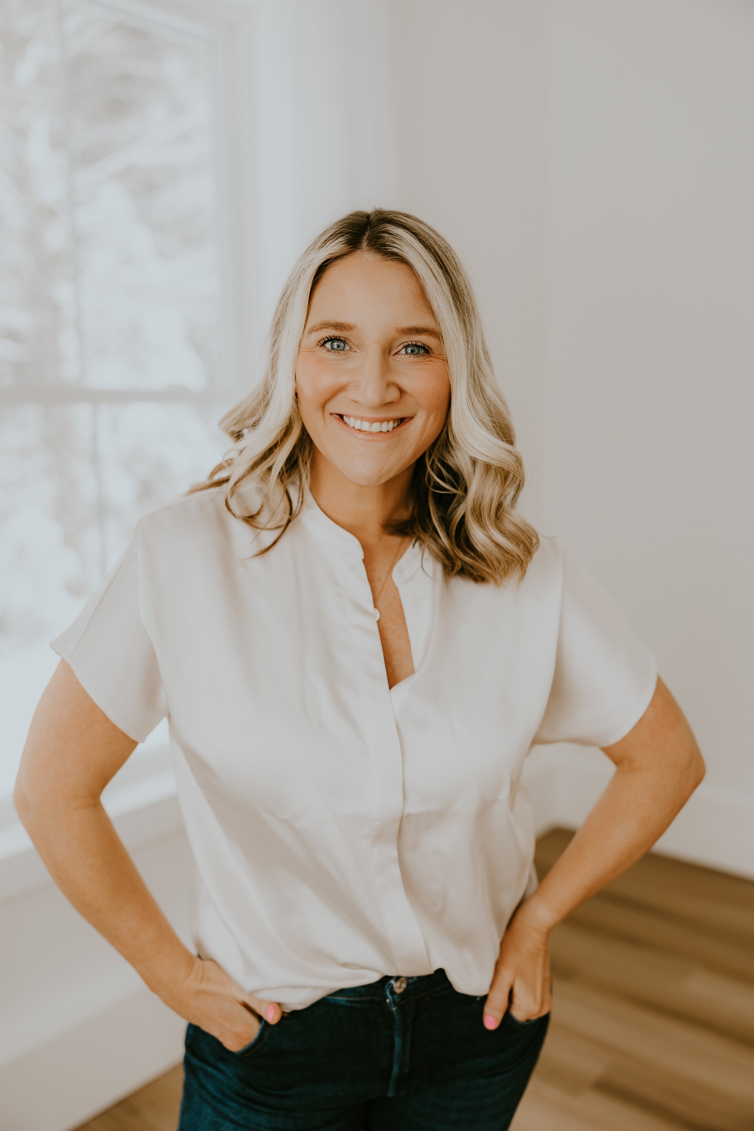 A smiling woman with blonde hair in loose waves, wearing a white blouse and dark jeans, standing indoors near a window with natural light.