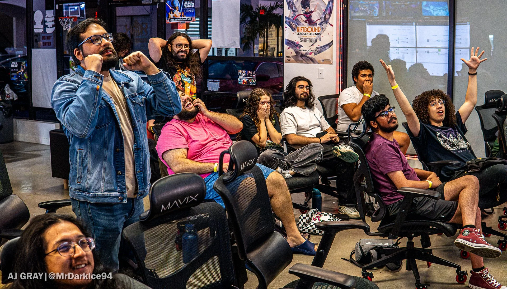 A group of people are seated and standing in a room, reacting to something off-camera. Expressions range from excitement to surprise, with several individuals raising their hands or covering their mouths. The room has posters on the walls, including one for "Riftbound" and a "Star Wars" themed sign. The setting appears to be a gaming or event space, with gaming chairs and posters visible.