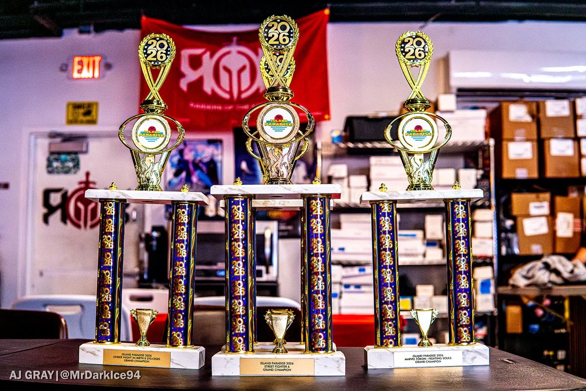 Three large trophies with gold, white, and blue colors and the year 2026 on top, displayed on a table in a room with shelves, boxes, and a red banner in the background. Each trophy has a plaque at the bottom indicating the event and year.