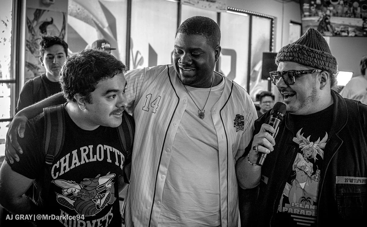 Three men at a social event, one holding a microphone, engaging in conversation, with others in the background.