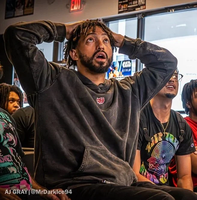 A man with dreadlocks and a beard, wearing a black hoodie, sits with his hands on his head, looking up with a surprised or shocked expression in a room with other people.