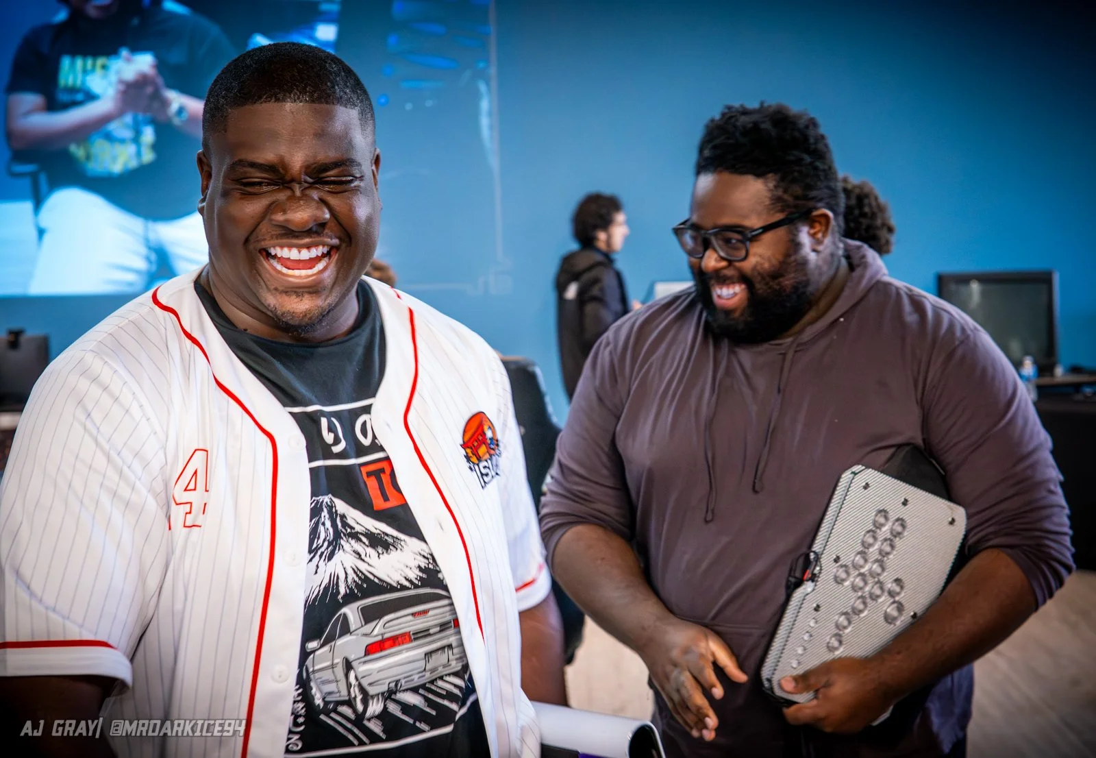 Two men are smiling and laughing in an indoor setting. The man on the left is wearing a baseball jersey and a graphic T-shirt, and the man on the right is wearing glasses and a hoodie, holding a device with a unique design. In the background, there are other people and computer monitors.