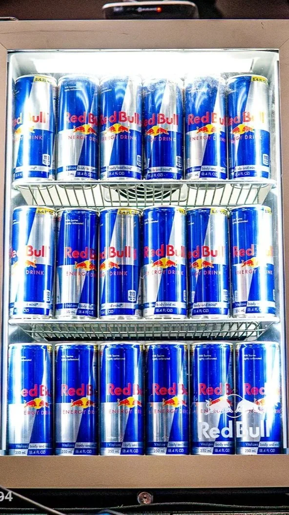 A refrigerated display case filled with rows of Red Bull energy drink cans, stacked in three shelves, featuring the recognizable blue and silver design with a red and yellow logo.