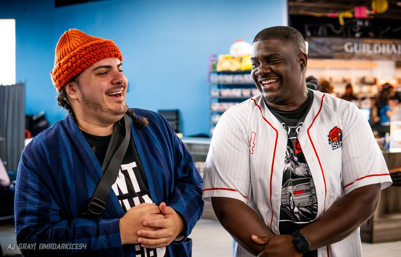 Two men smiling and talking inside a retail store, one wearing an orange knit beanie and a blue jacket, the other wearing a white sports jersey with red accents.