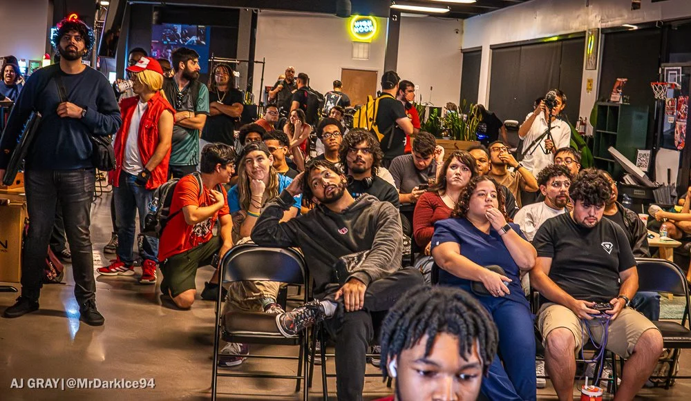 A large group of people seated and standing in a gaming or event space, with many focused on an unseen speaker or screen. The room has dark walls, plants, and arcade/game equipment visible in the background.