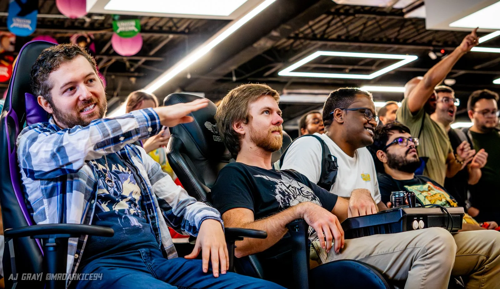A group of people attending a gaming or tech event, sitting and standing in a crowded indoor space, with some showing excitement and others focused on the event.