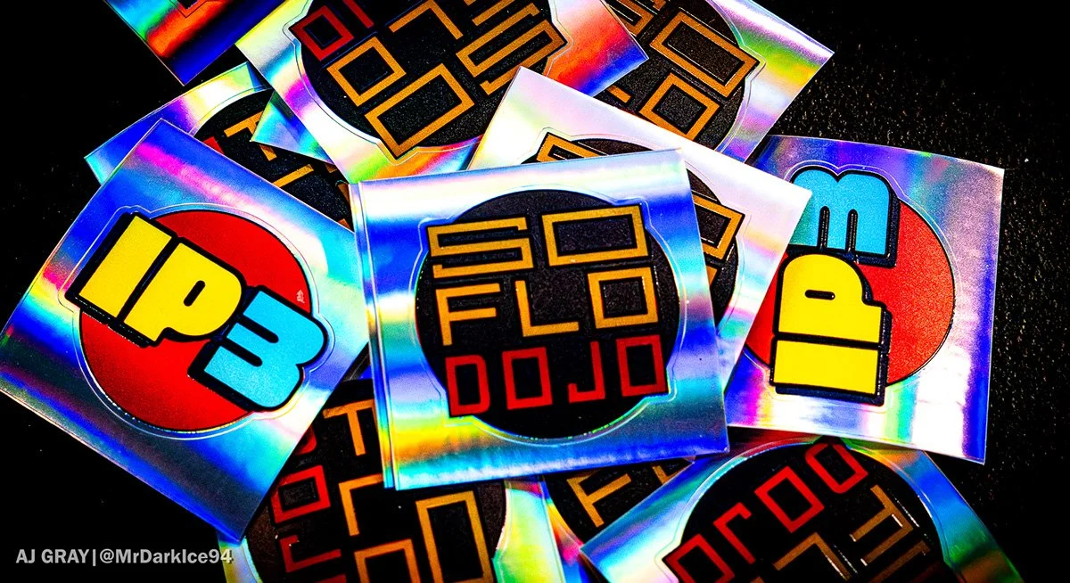 Colorful holographic stickers with black backgrounds and bold, blocky text. Some stickers say "IP3" with a red circle behind the text, and others say "SOFLO DOJO" with a black circle background.