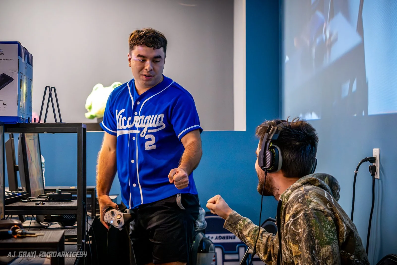 Two young men in a room, one standing and the other sitting with gaming headphones, fist bumping, with gaming and electronic equipment around them.