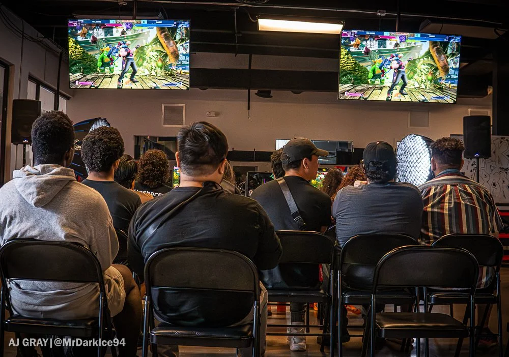 Audience watching a video game on two large screens in a gaming event or arcade, with players competing on the screens.