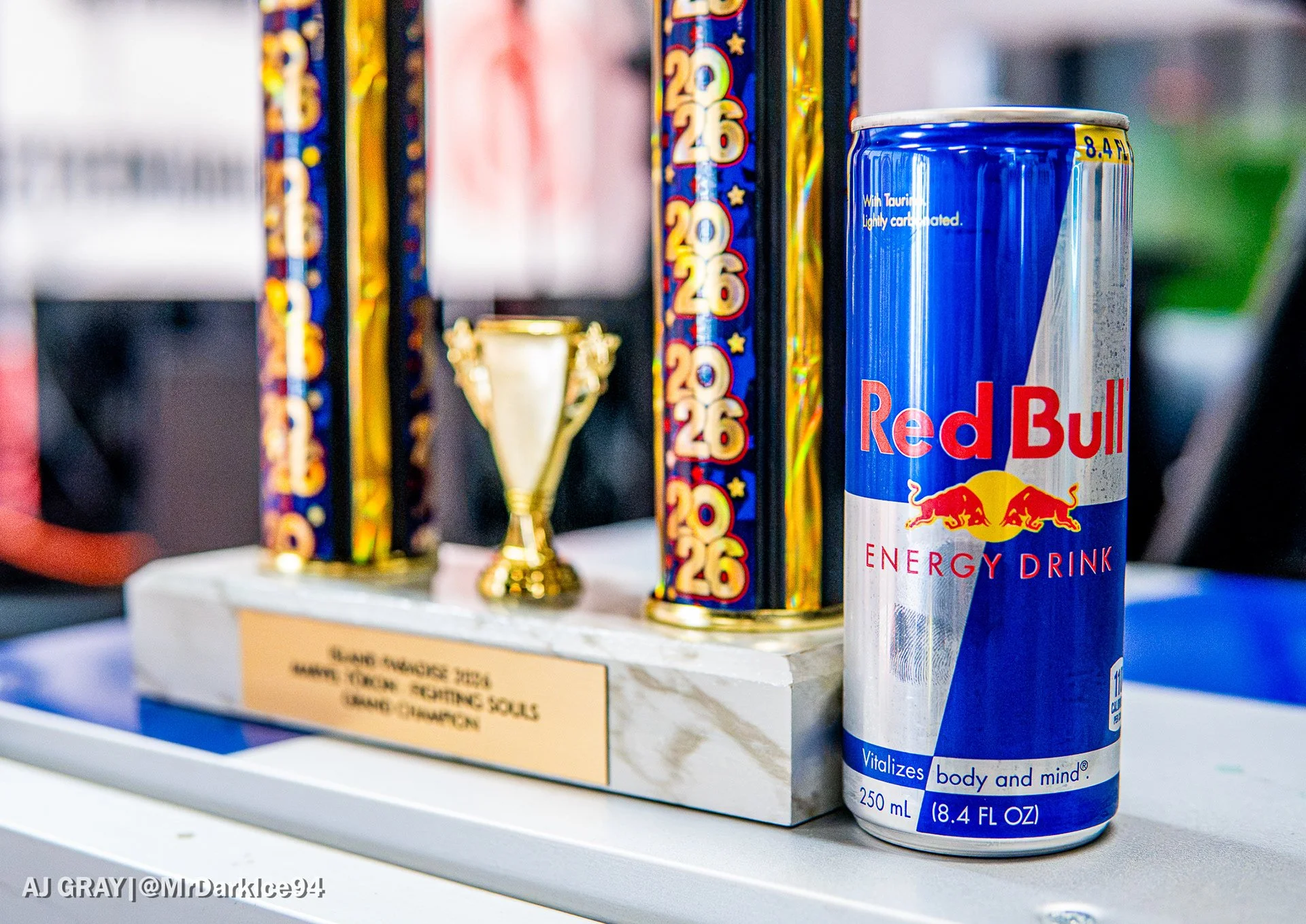 A can of Red Bull energy drink placed on a white surface in front of a row of colorful fireworks and a small gold trophy. The can is blue and silver with red and yellow accents, and the fireworks have red, yellow, blue, and black colors with star and number patterns.