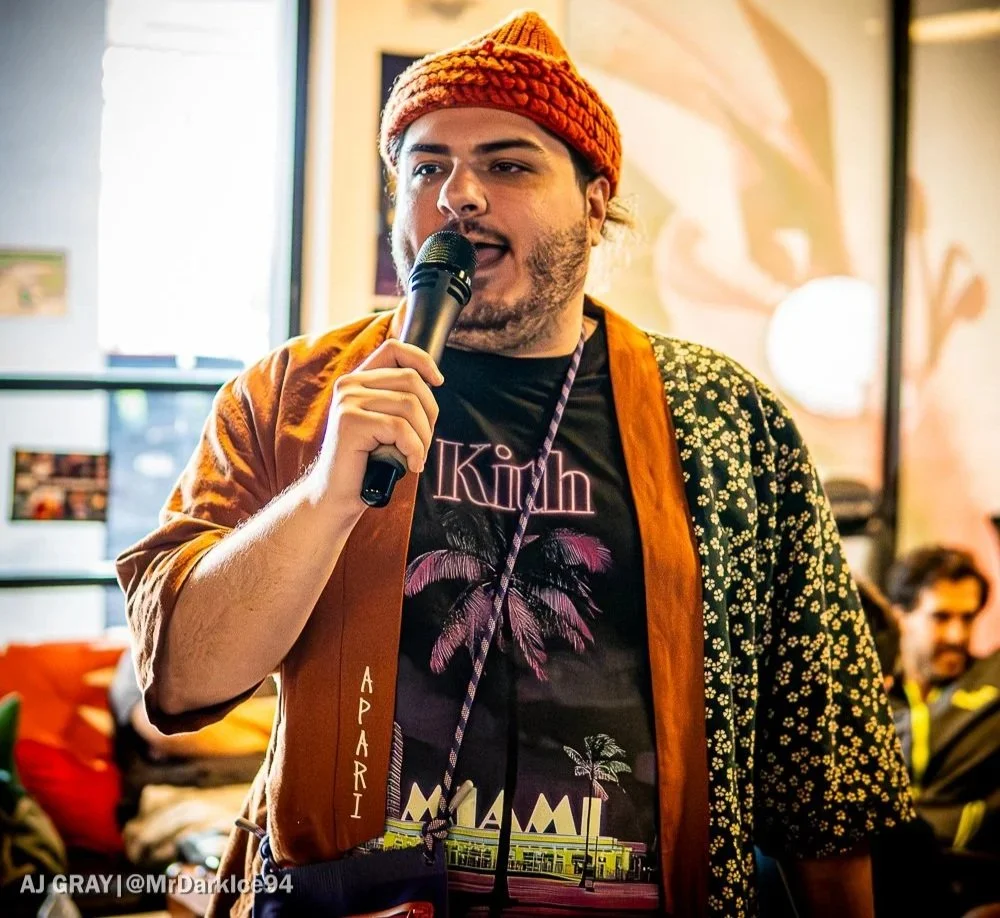 A man in a beanie hat holding a microphone, speaking at an indoor event with warm lighting and people in the background.