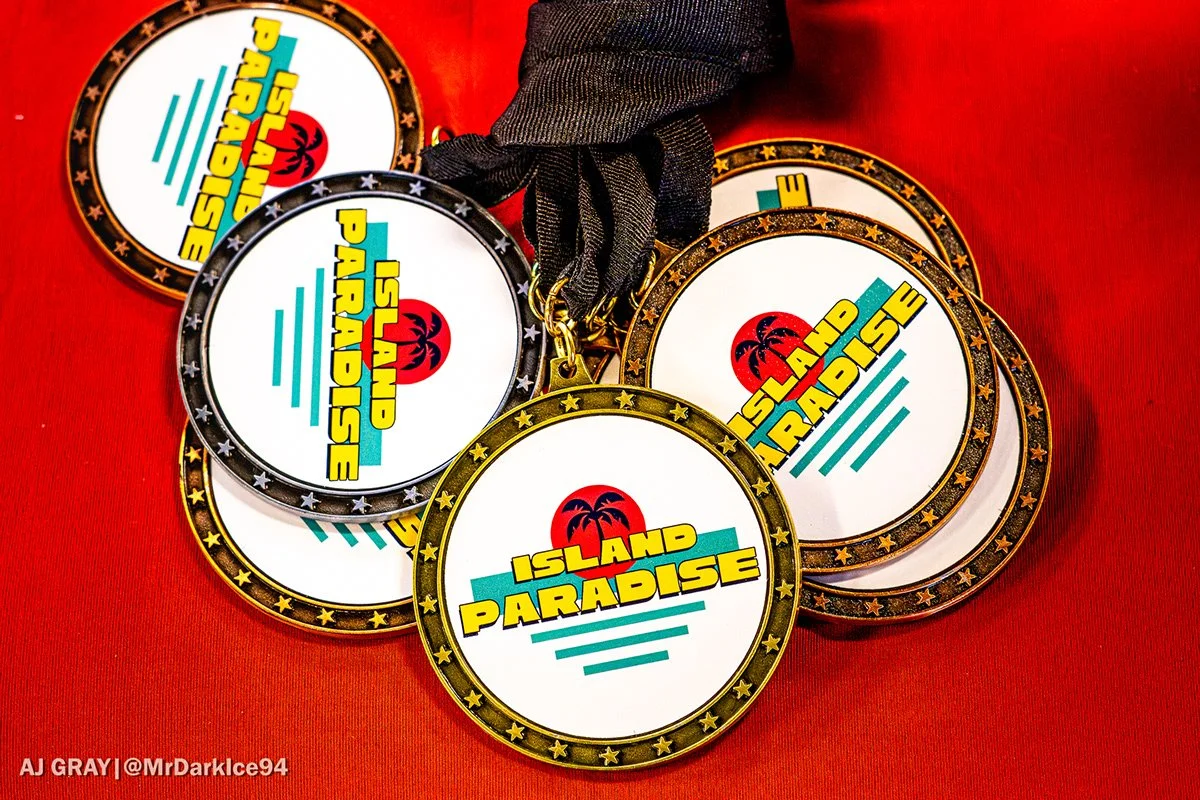 Several medals with the inscription 'Island Paradise' and a palm tree logo, placed on a red surface. The medals have a gold, black, or silver border with star designs, and are attached to ribbons.
