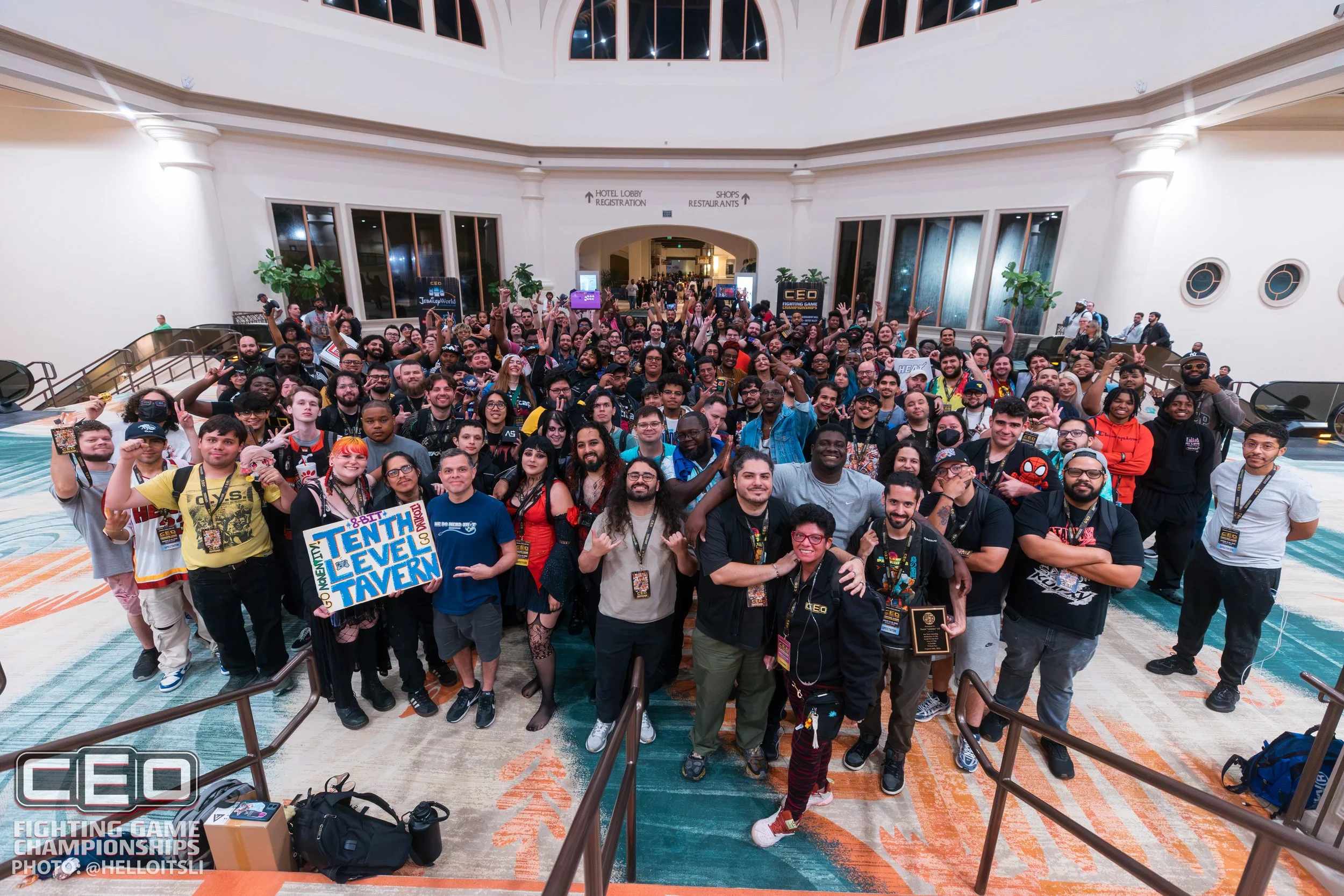 A large group of people gathered in a spacious, well-lit indoor area, smiling and posing for a group photo at a gaming or comic convention.