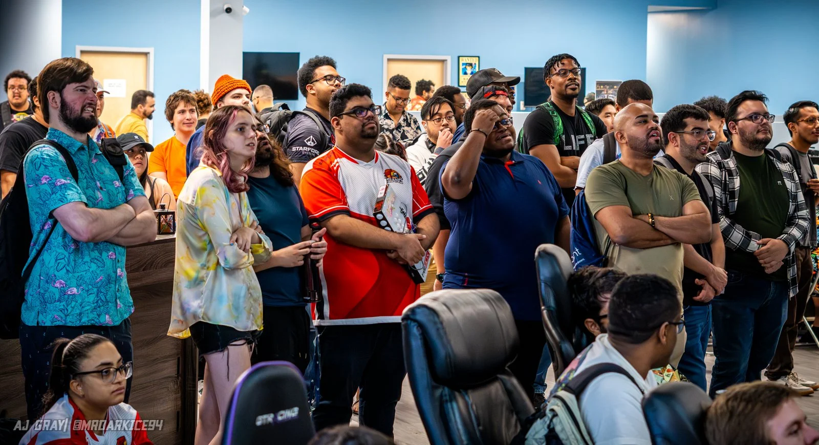 A group of diverse people standing and watching attentively at an event, with some crossing their arms and others holding items, in a room with blue walls and framed pictures.