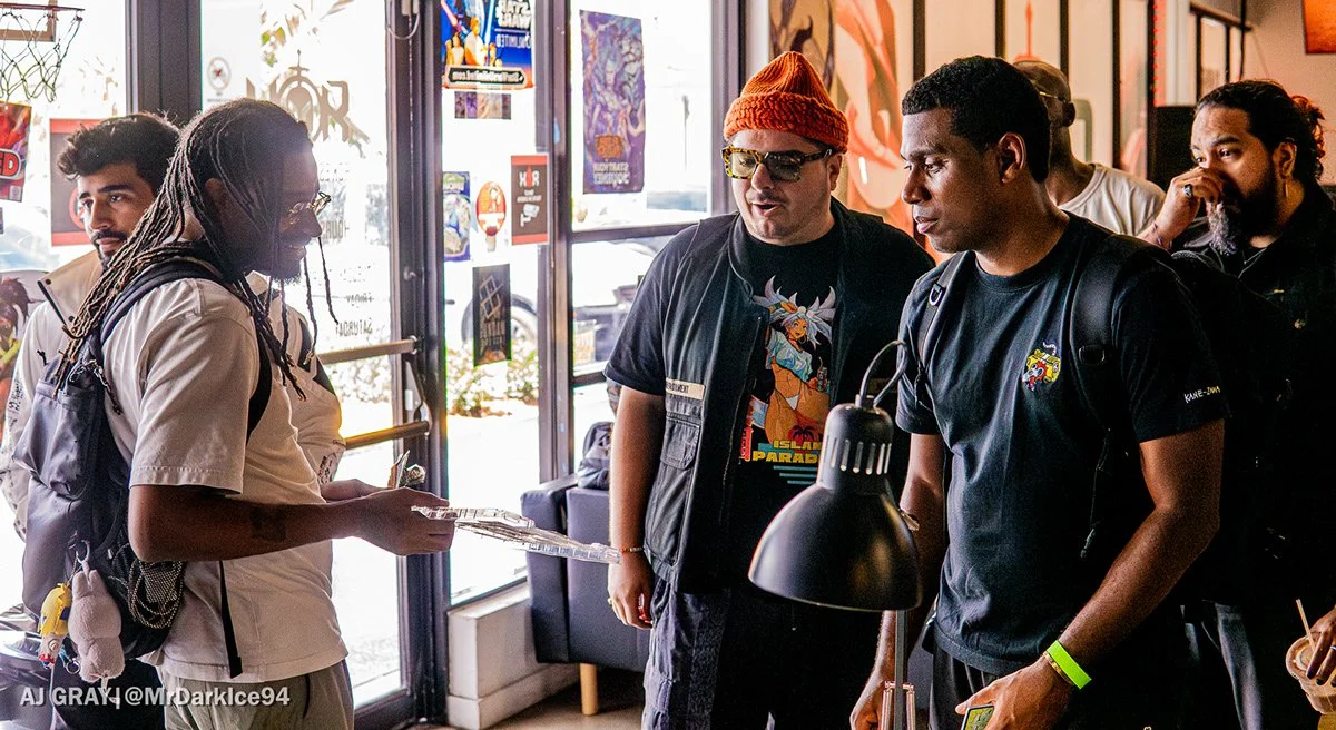 People gathered inside a cafe or restaurant, engaging in conversation. There are five individuals, each with distinctive styles, standing near a table with a black lamp. The background shows posters and artwork on the walls and large windows letting in natural light.