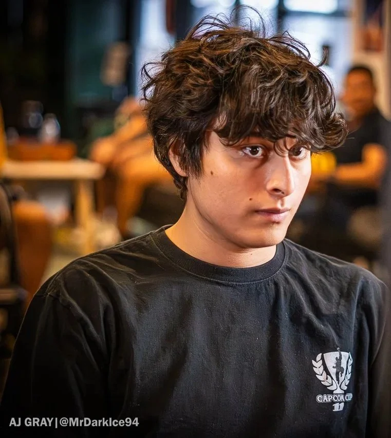 A young person with messy, curly brown hair wearing a black T-shirt with a Capcom Cup logo on it, sitting indoors with blurred people in the background.