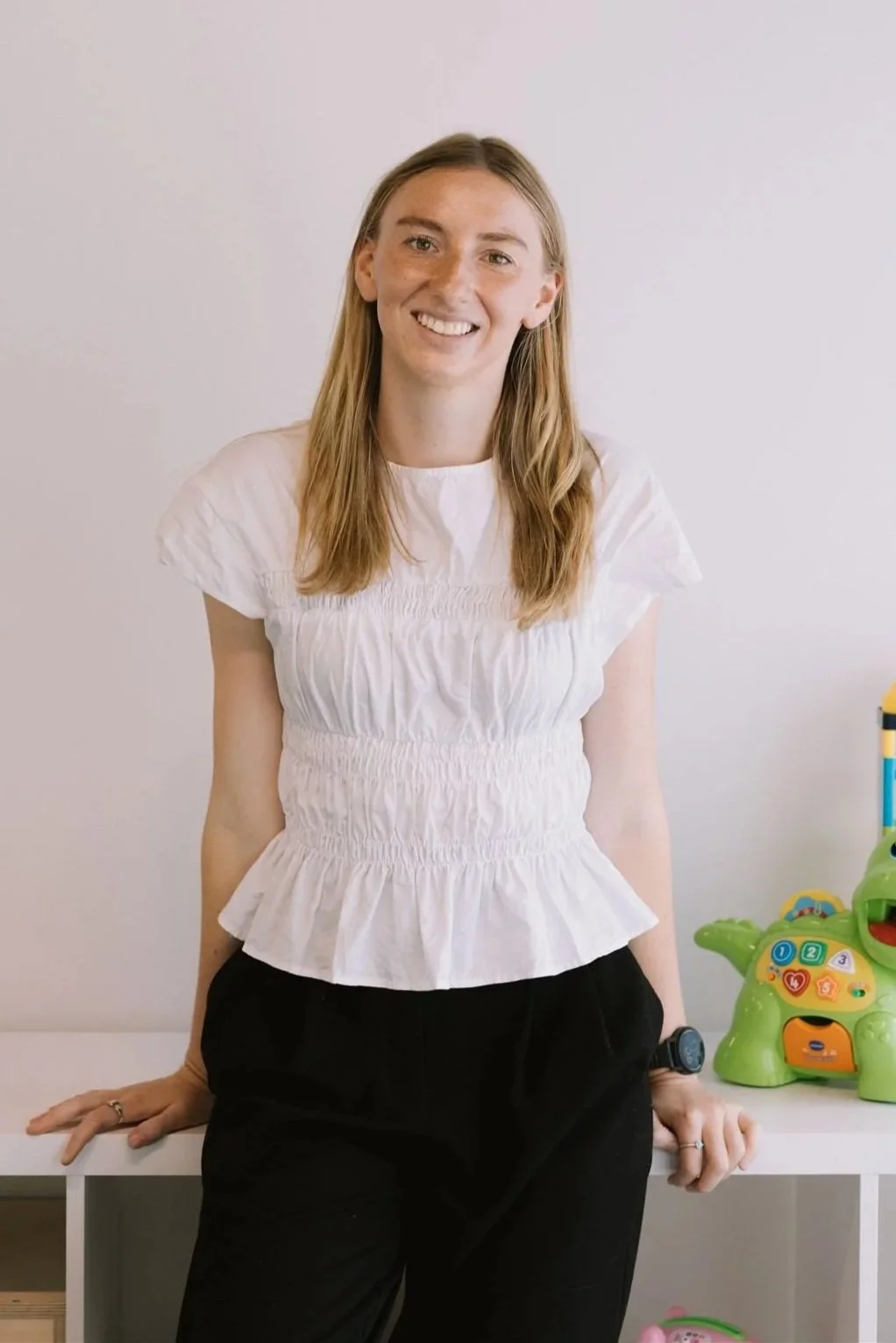 A woman with long blonde hair, wearing a white top and black pants, standing and leaning on a white table with her left hand, smiling at the camera. There is a colorful toy dinosaur on the right side of the table.