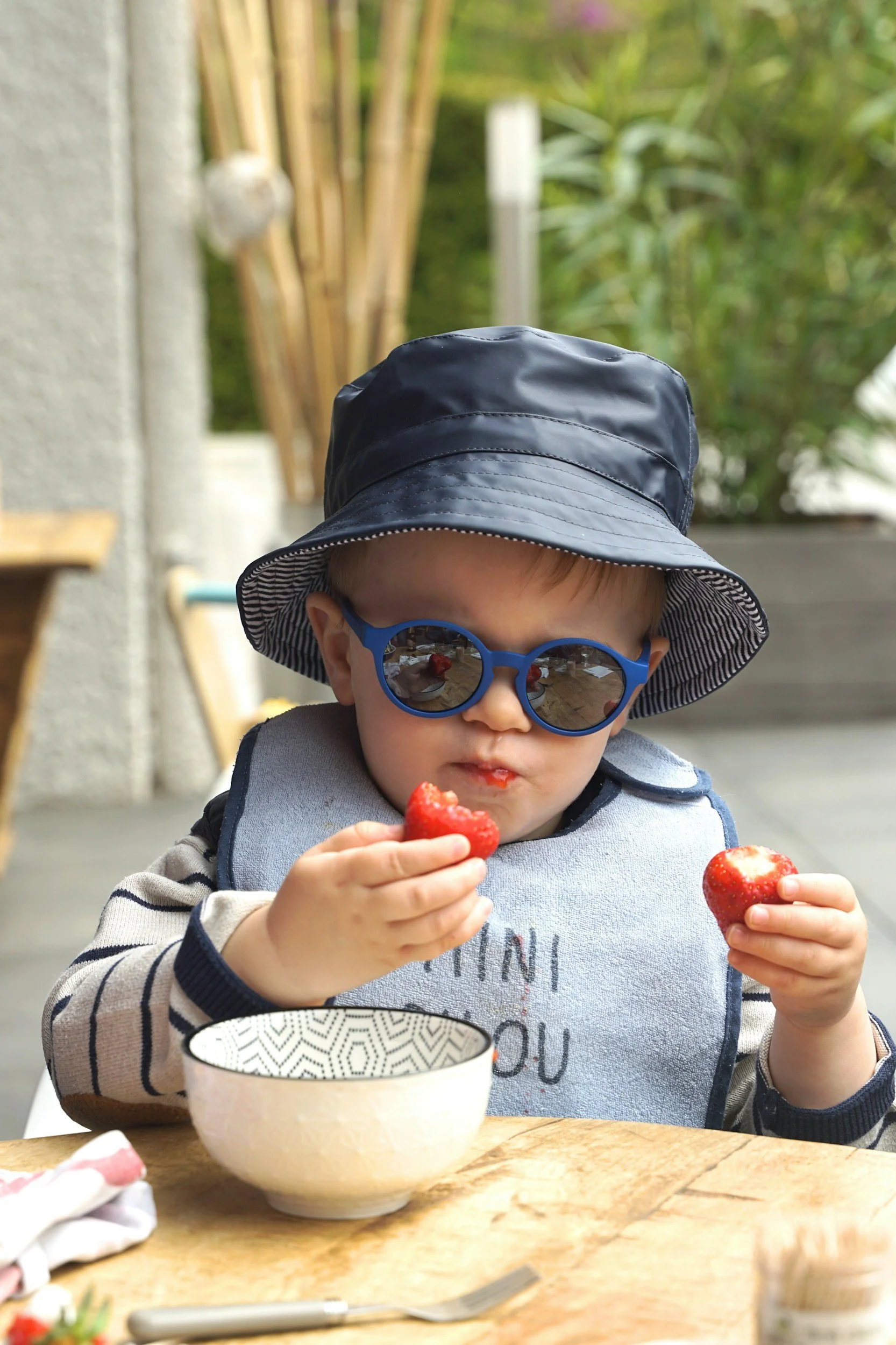 A young child wearing a striped long-sleeve shirt, blue sunglasses, and a black sun hat, sitting at a wooden table outdoors, eating strawberries from a bowl.