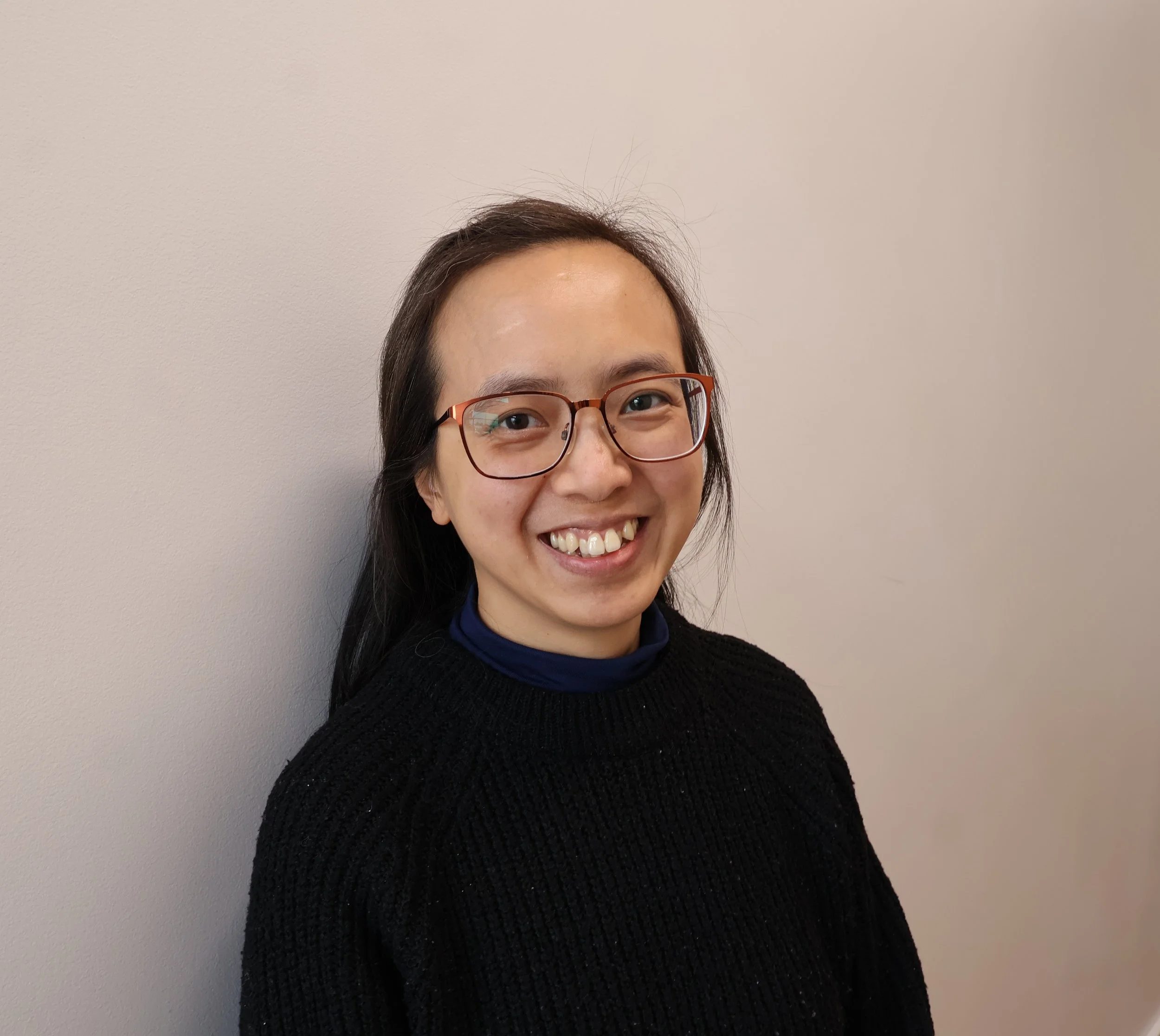 Close-up of a smiling woman with glasses, wearing a dark sweater, standing in front of a plain wall.