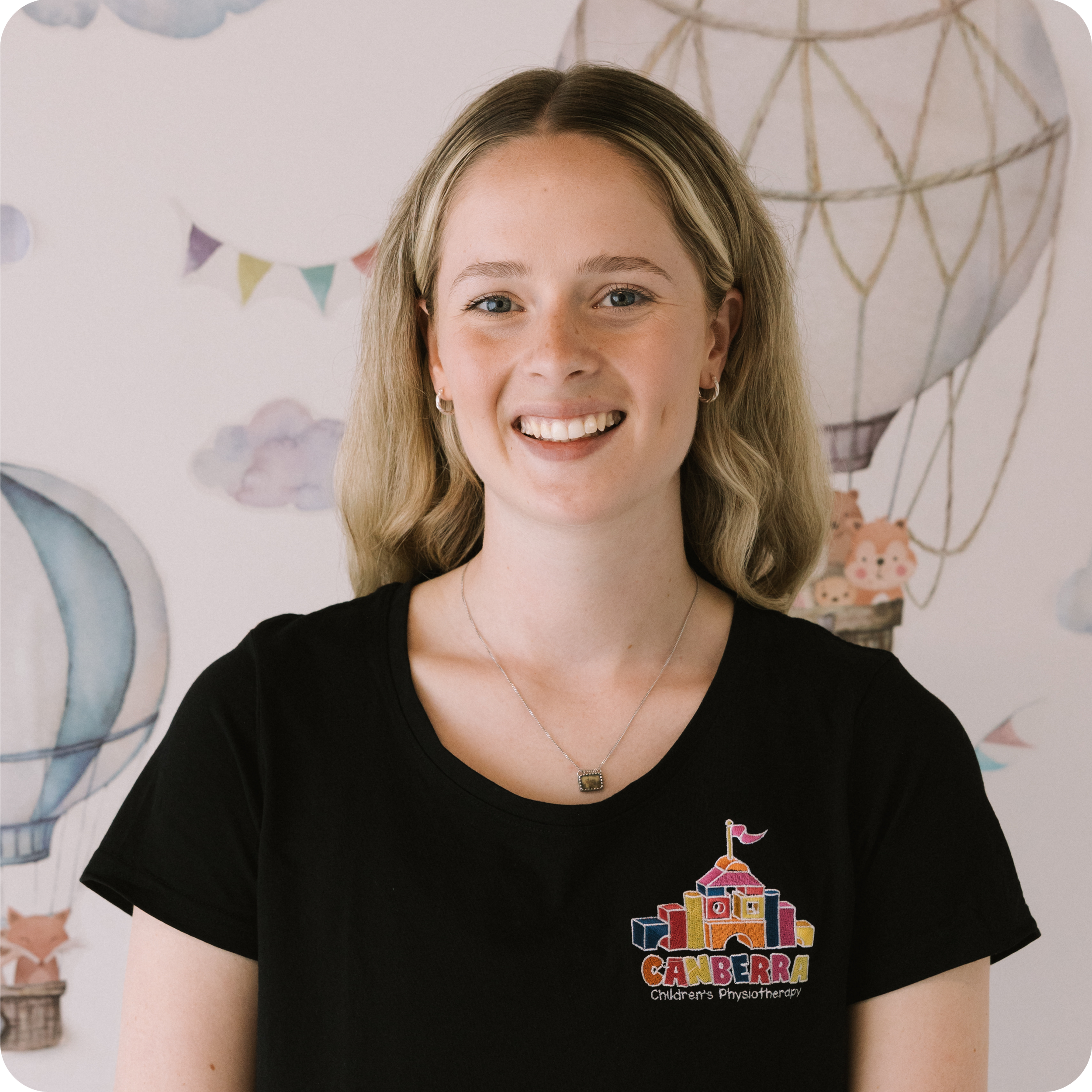Close-up of a smiling young woman with blonde hair wearing a black shirt and a necklace, standing in front of a wall with colorful decorations, including a hot air balloon and bunting, at a children's physiotherapy center in Canberra.