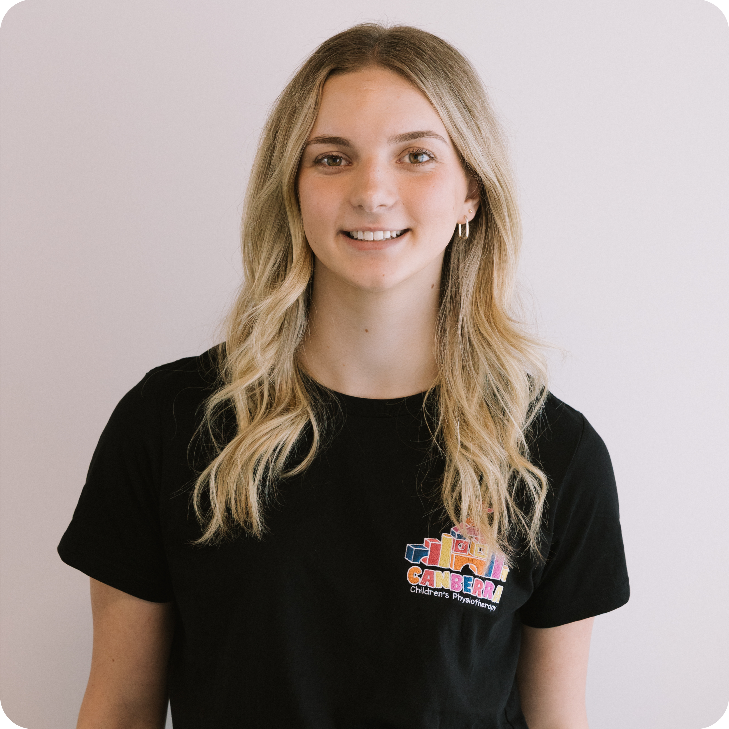 Close-up of a young woman with long blonde hair wearing a black t-shirt with a colorful logo for Children's Physiotherapy. The photo is taken against a plain light background.