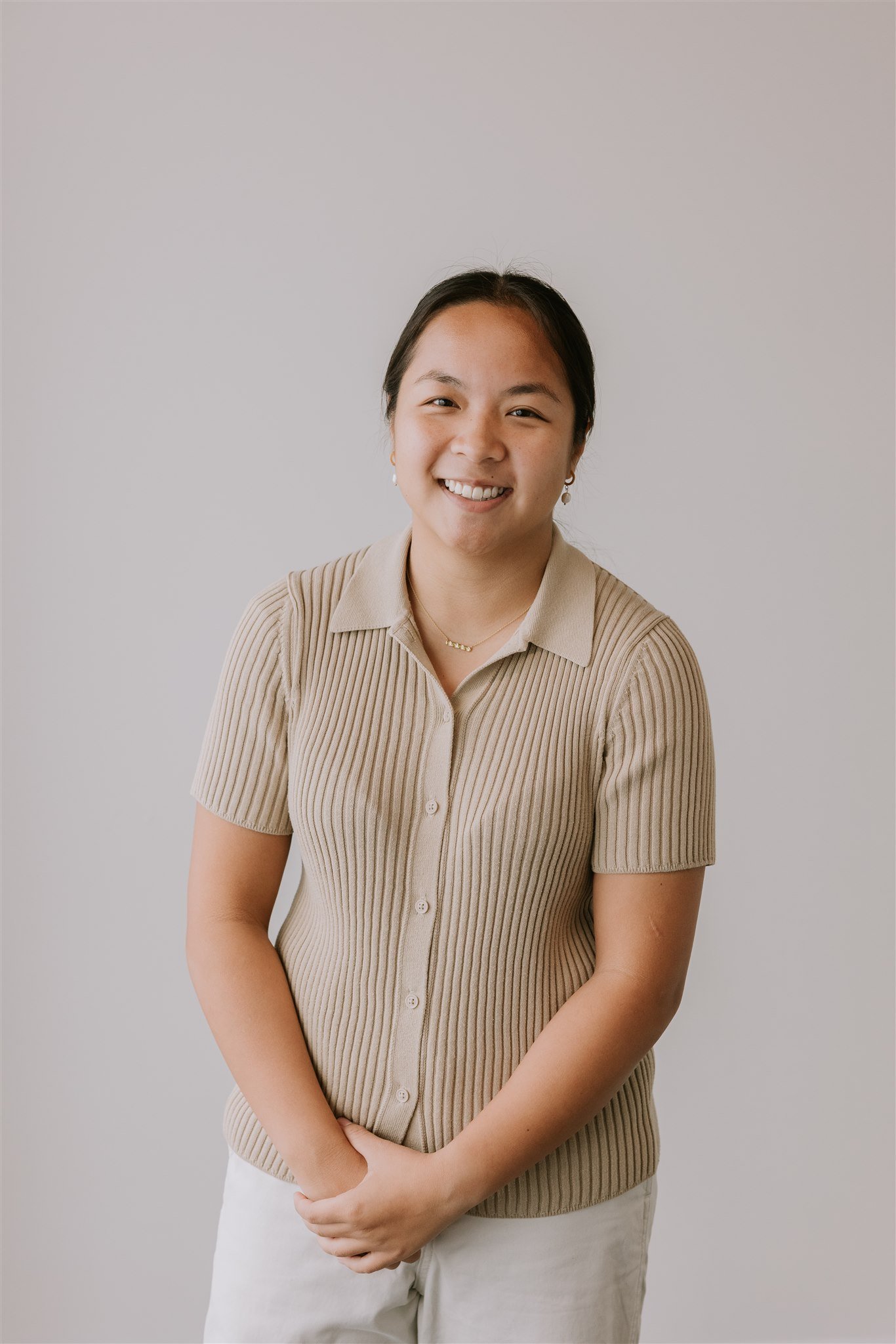 Lauren Ngyuen, PT - woman with dark hair tied back, smiling, wearing a beige, short-sleeve, button-up, ribbed shirt and jewelry, standing against a plain light background.