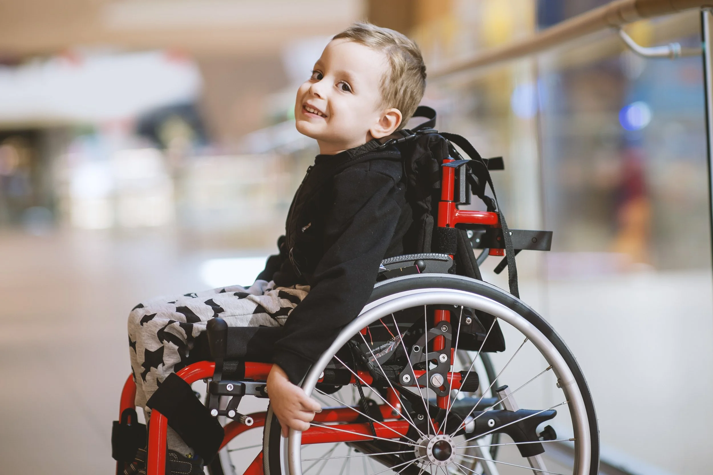 A smiling young boy in a wheelchair in a brightly lit indoor setting.
