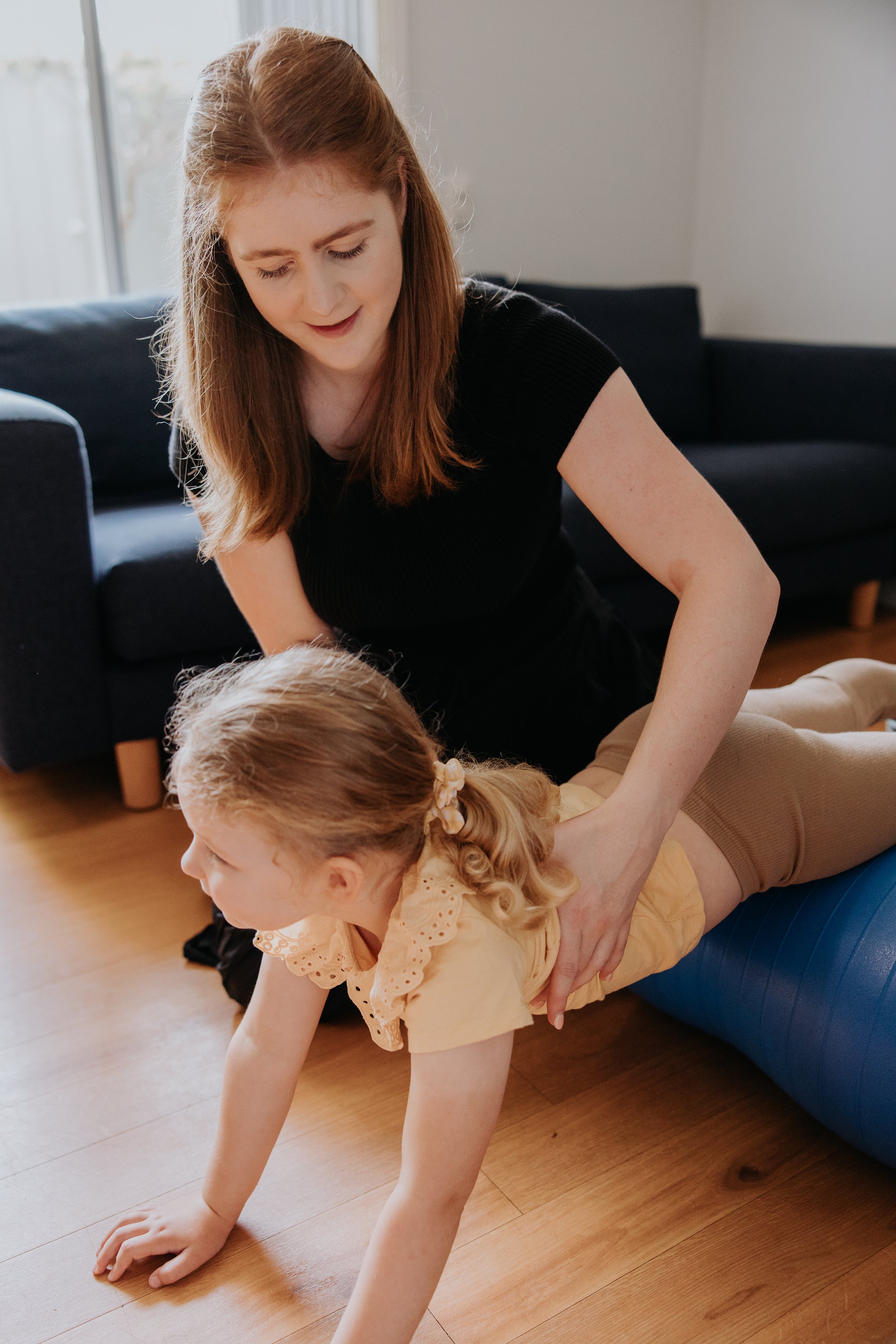 A woman with red hair helps a young girl with curly blonde hair do a back stretch on a wooden floor in a living room with natural light.
