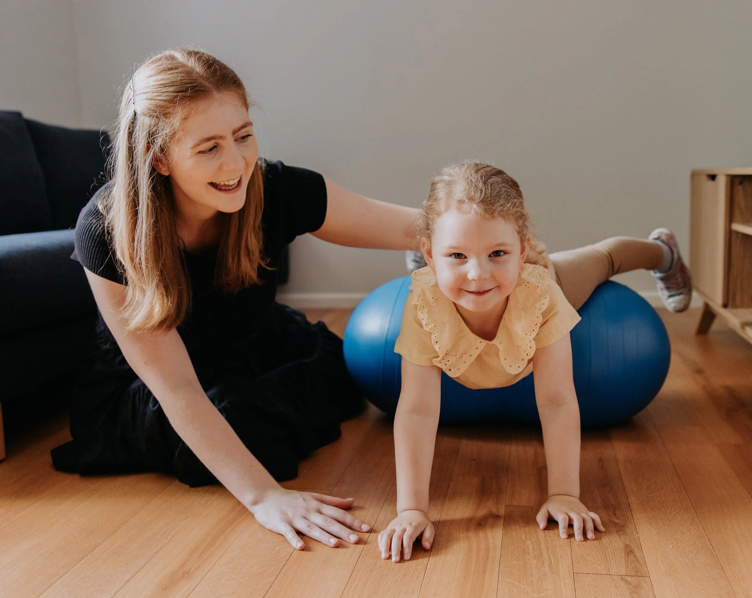 A woman helping a young girl do a tummy exercise on a blue exercise ball in a living room.